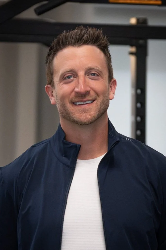 A smiling man with short brown hair and light stubble, wearing a navy blue jacket over a white shirt, standing in a gym or fitness studio.