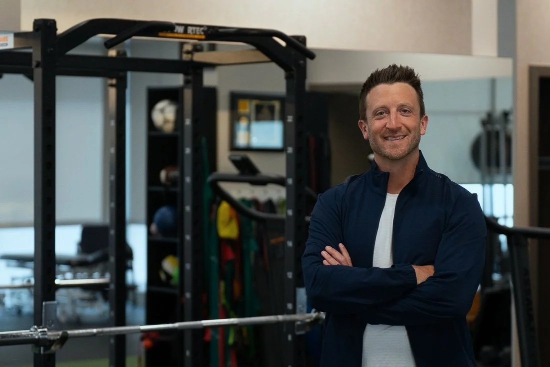 A man with short brown hair and a beard smiling, standing with arms crossed in a gym, with workout equipment in the background.