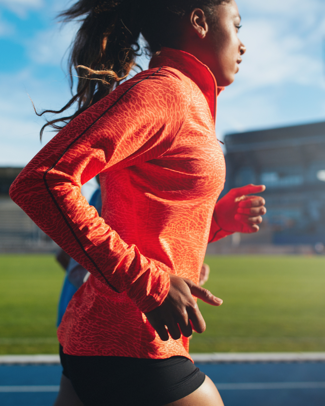 A woman running on a track, wearing a bright orange athletic jacket and black shorts, with her hair flowing behind her.