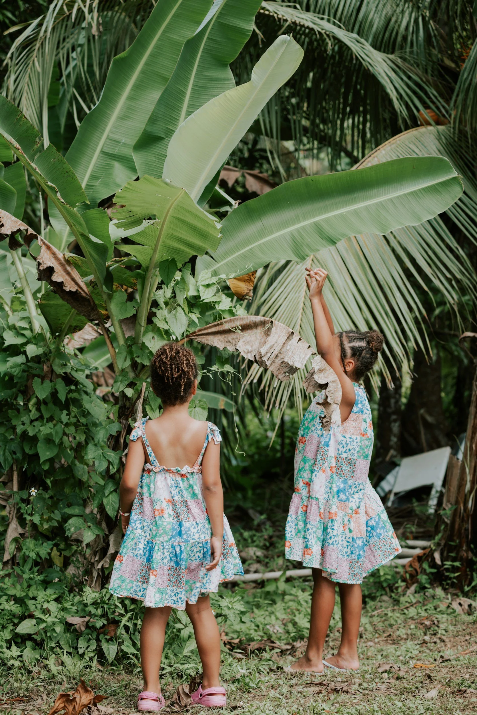 Two young girls in matching floral dresses playing in a lush tropical garden surrounded by large green banana and palm leaves.