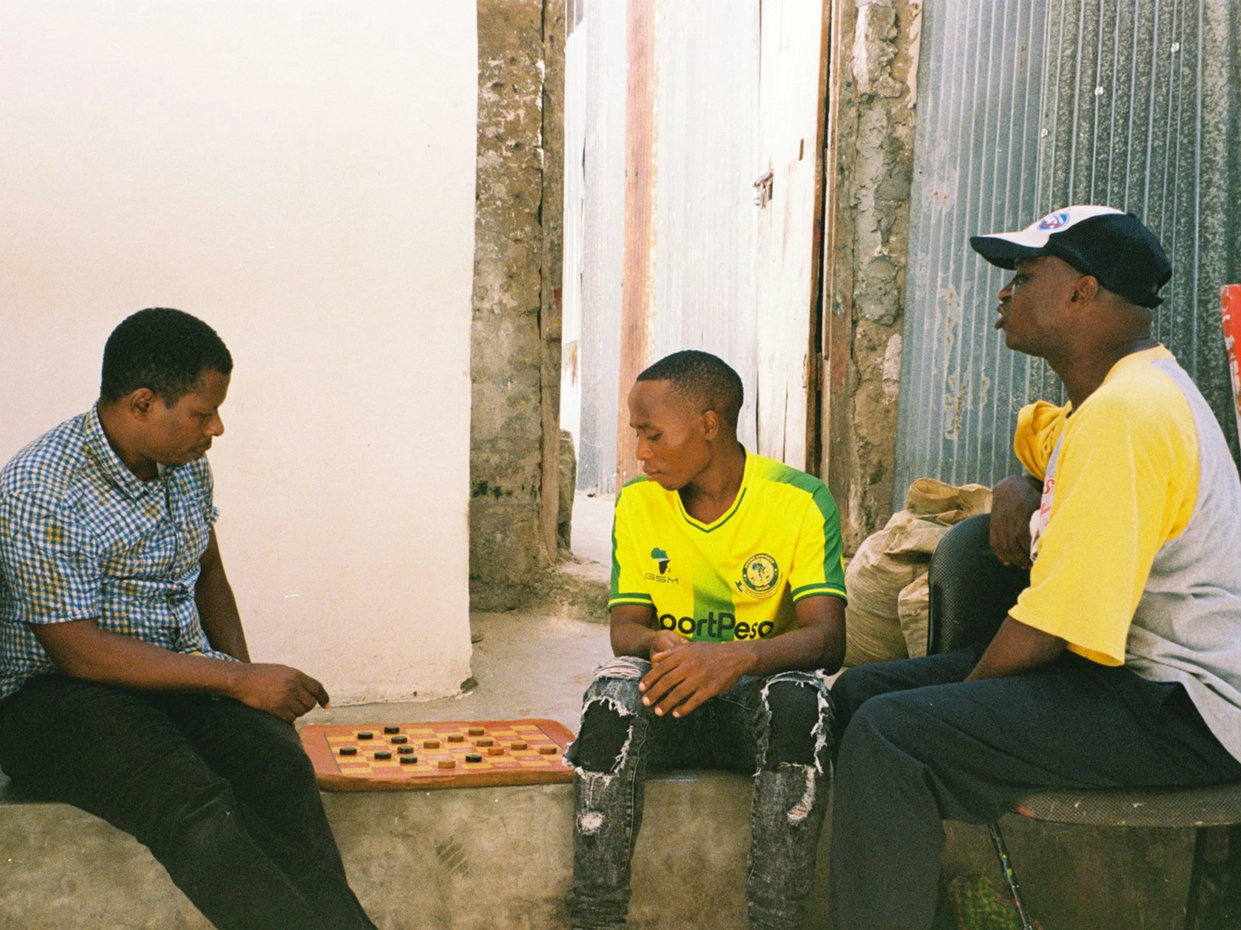 Three young men sitting and playing a game of checkers in a room with unfinished walls and a doorway in the background.