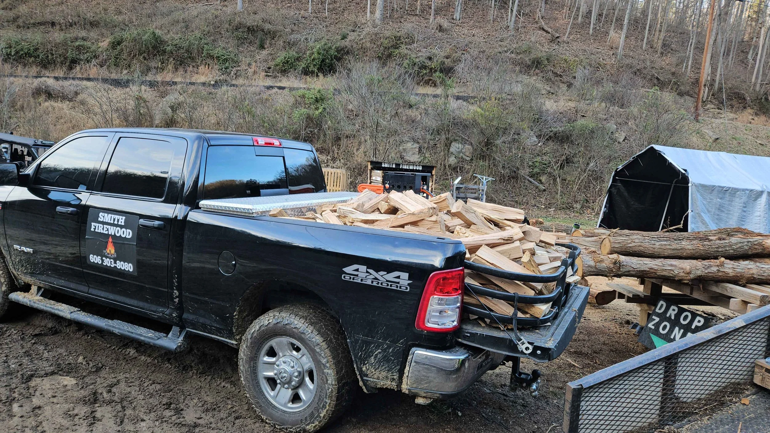 A black pickup truck filled with chopped firewood, parked on muddy ground near a hillside. There is a sign that says 'Drop Zone' near the truck, and a tent is visible in the background.