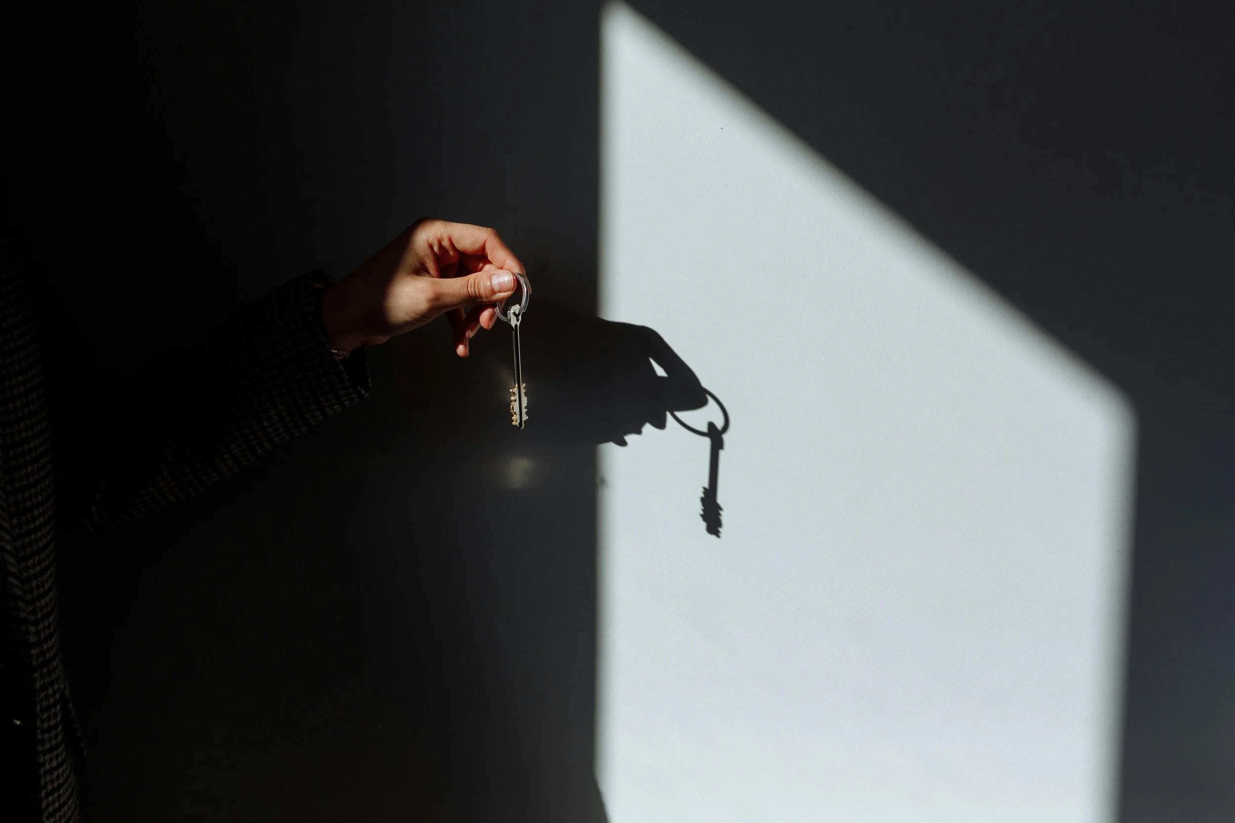 A person's hand holding a key with a shadow cast on a wall, lit by sunlight from a window.