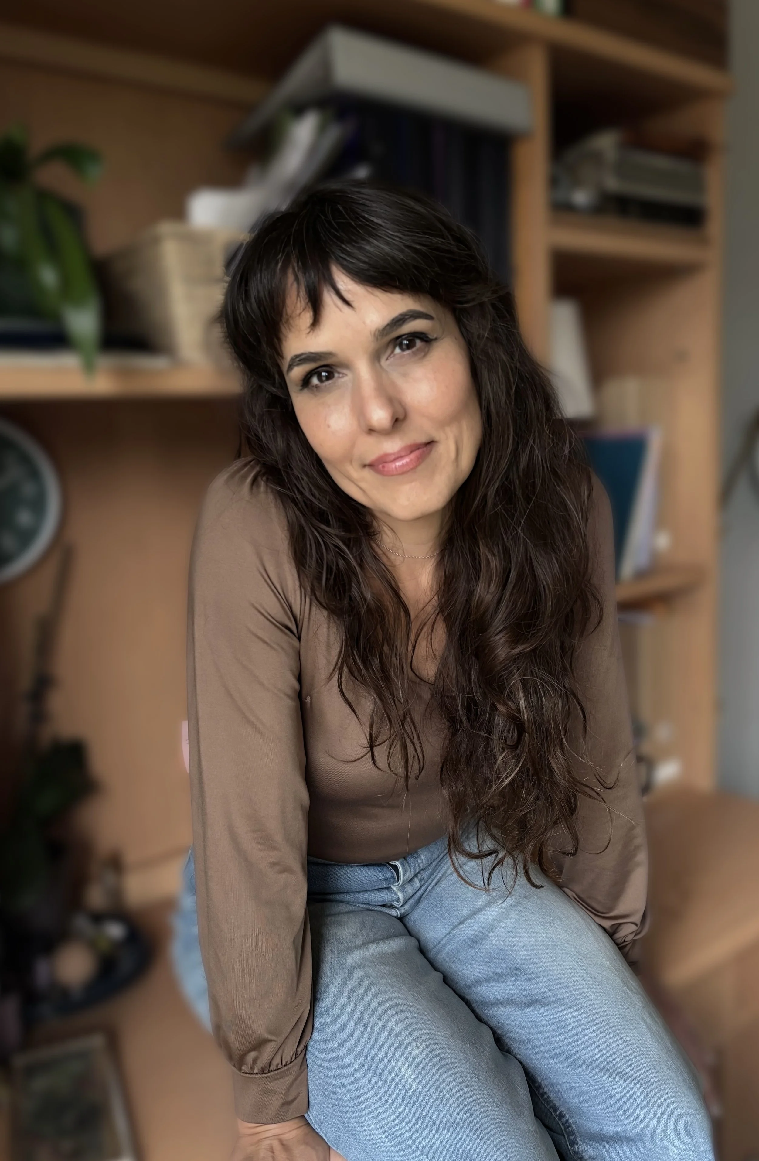Mary Bradford,  with long dark wavy hair smiling at the camera, sitting on a wooden surface with bookshelves and plants in the background, ready to work on your health problems.