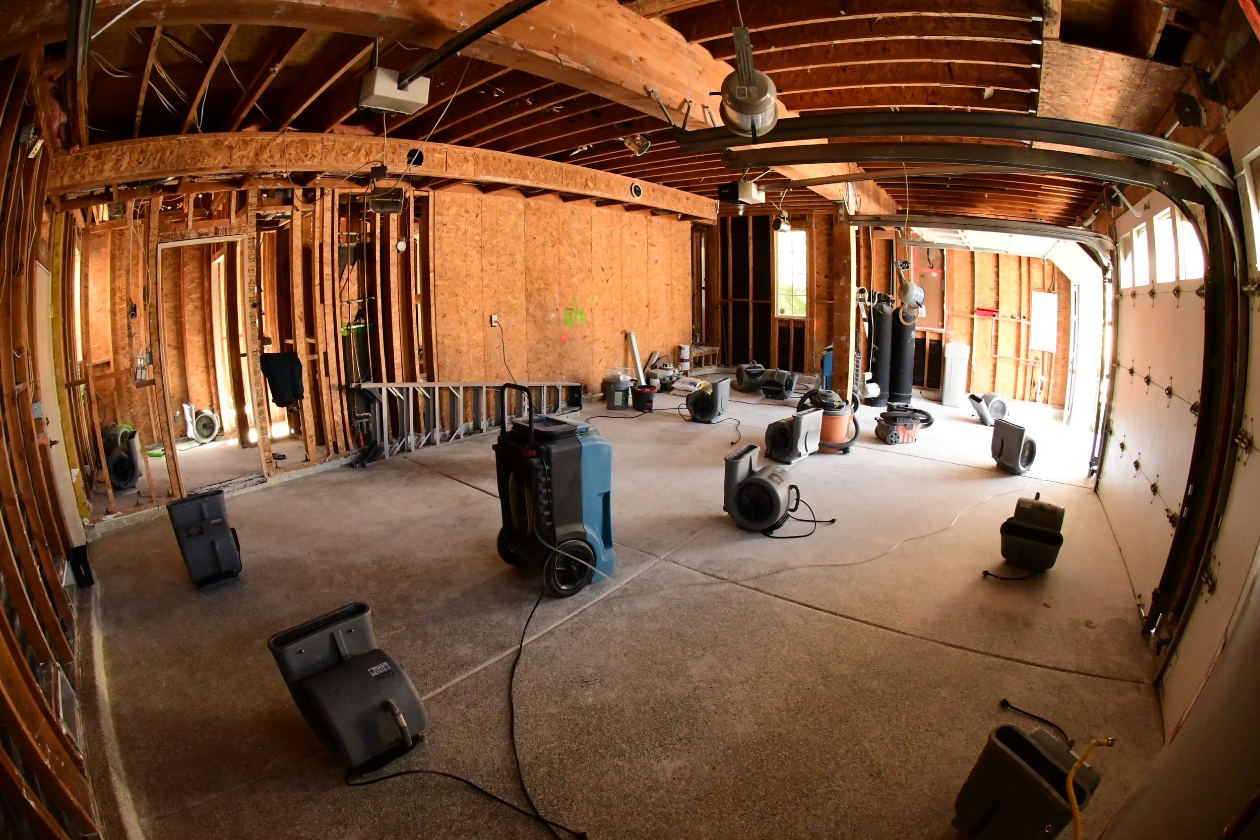 Interior view of a garage under construction with unfinished wooden framing on walls, open garage door, and various portable dehumidifiers scattered on the carpeted floor.
