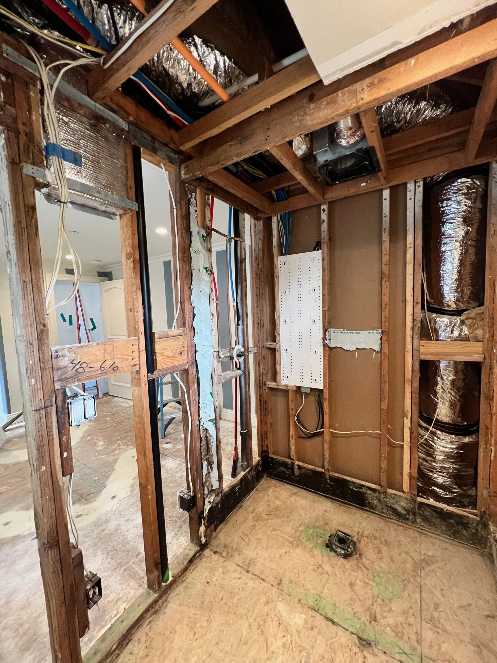 Interior view of a room under renovation with exposed wooden framing, electrical wiring, and insulation. The floor is unfinished, and there is a white electrical panel or junction box on the wall.