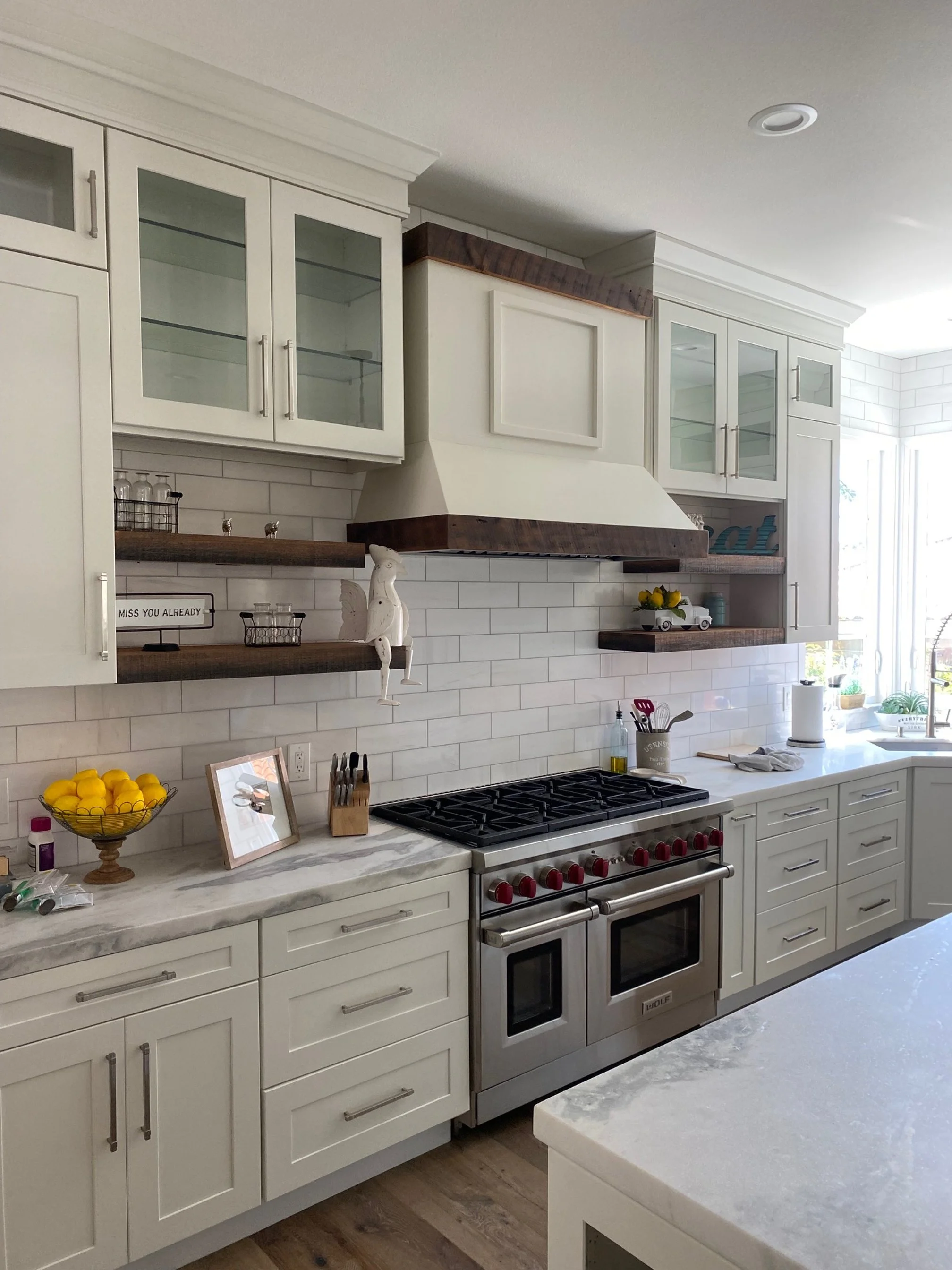 Modern kitchen with white cabinets, marble countertops, a stainless steel double oven, open wooden shelves, white tile backsplash, and a large window letting in natural light.