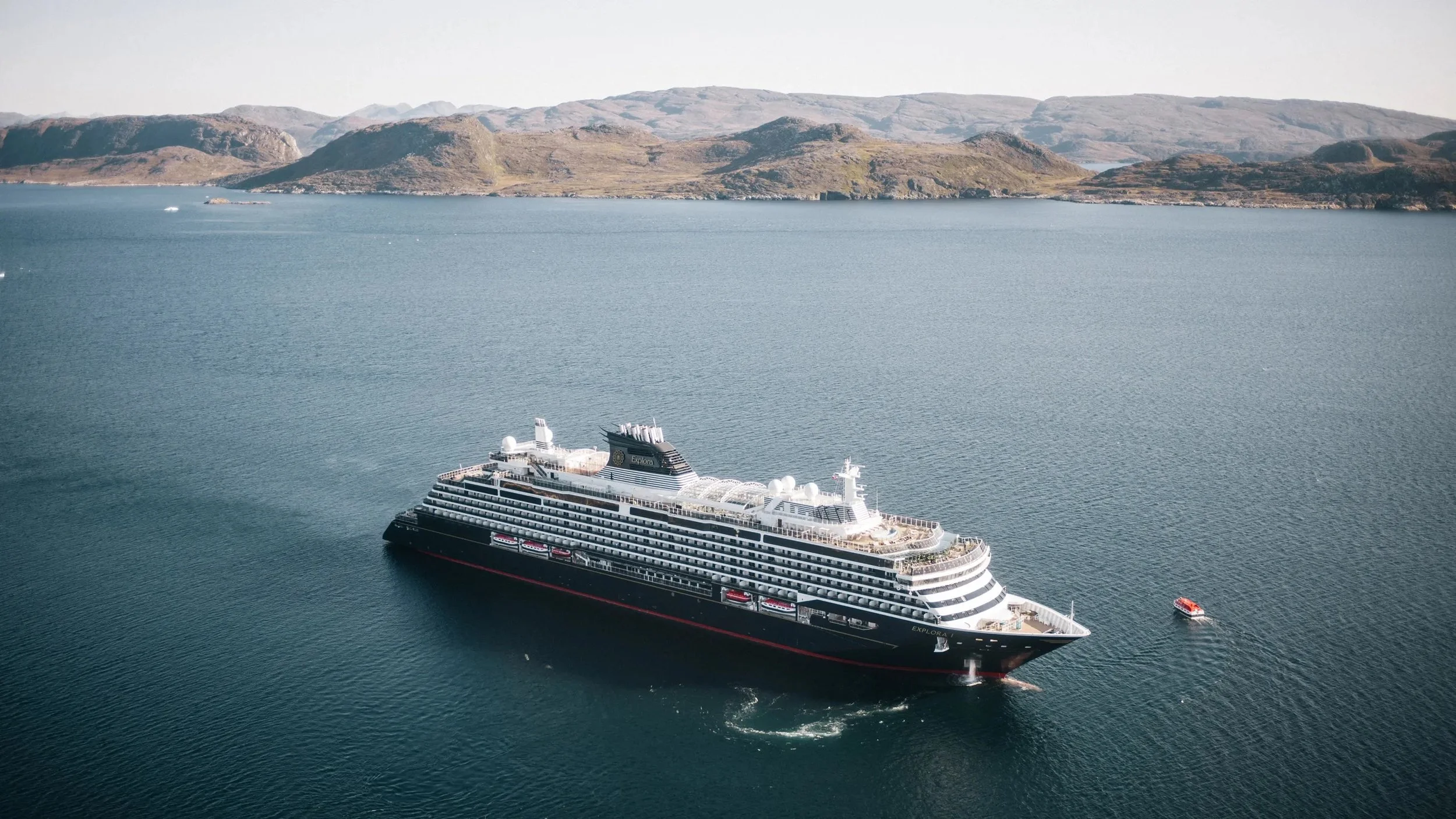 A large cruise ship sailing in a body of water with hills in the background. The cruise ship is black and white with multiple decks and a red stripe along the bottom.