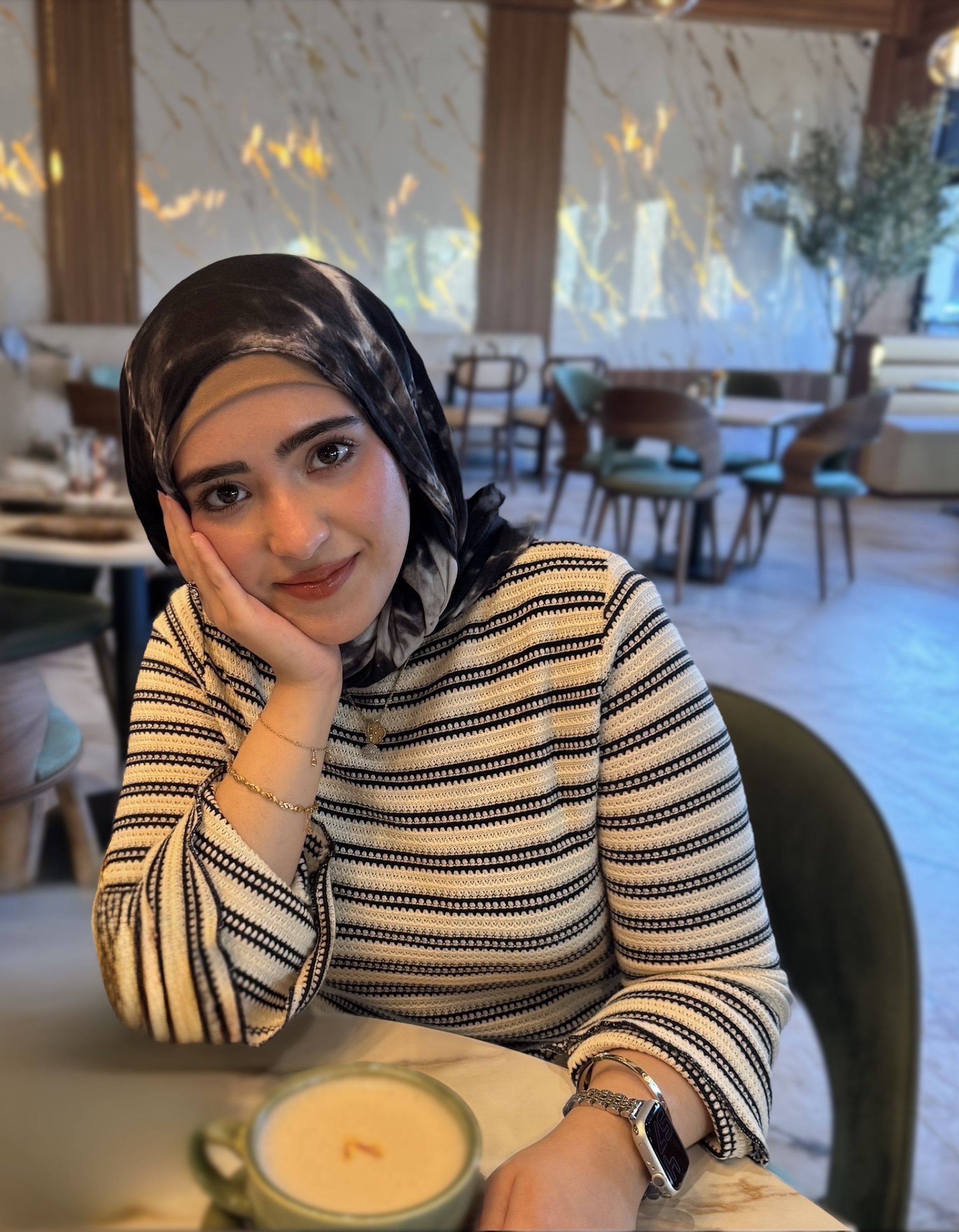 Muslim Therapist / Islamic Psychologist resting her face on her hand, with a mug of coffee on the table in front of her. The background shows a wooden wall with abstract yellow and gold leaf designs, tables, chairs, and a window with a view outside.