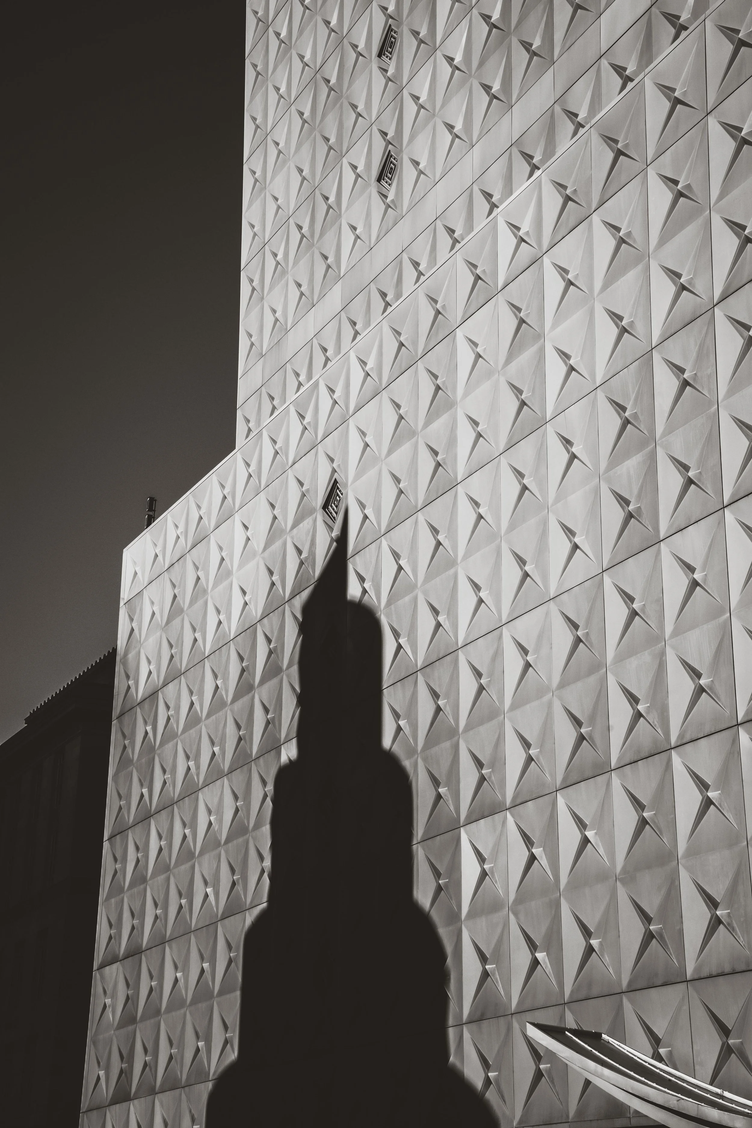 Shadow of a person holding up a smartphone, cast on a modern building with a geometric patterned facade.