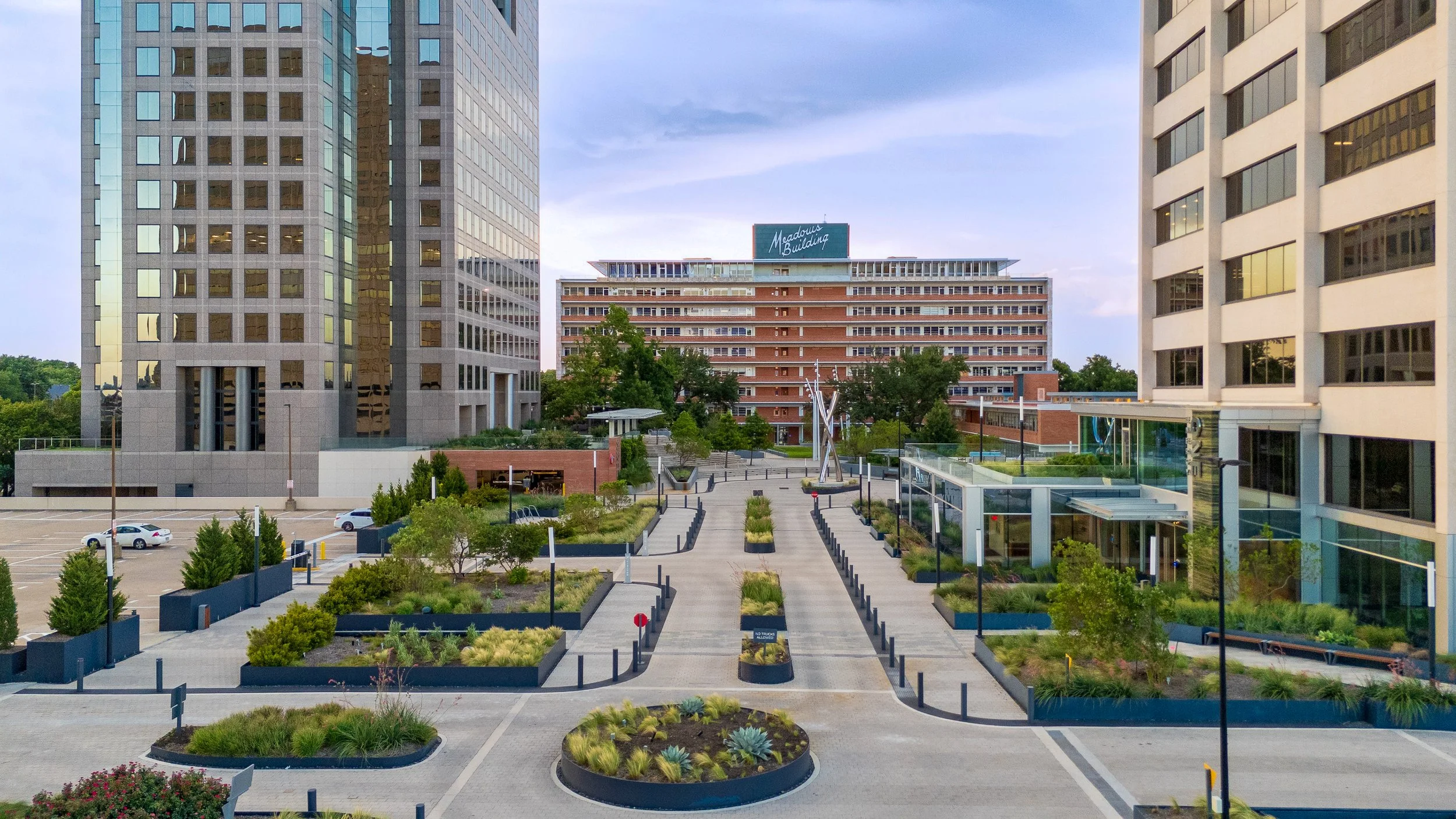 View of a modern cityscape with tall office buildings, landscaped pathways, small trees, benches, and parking lot in the foreground. The central building has a sign that says 'Meadow's Building'.