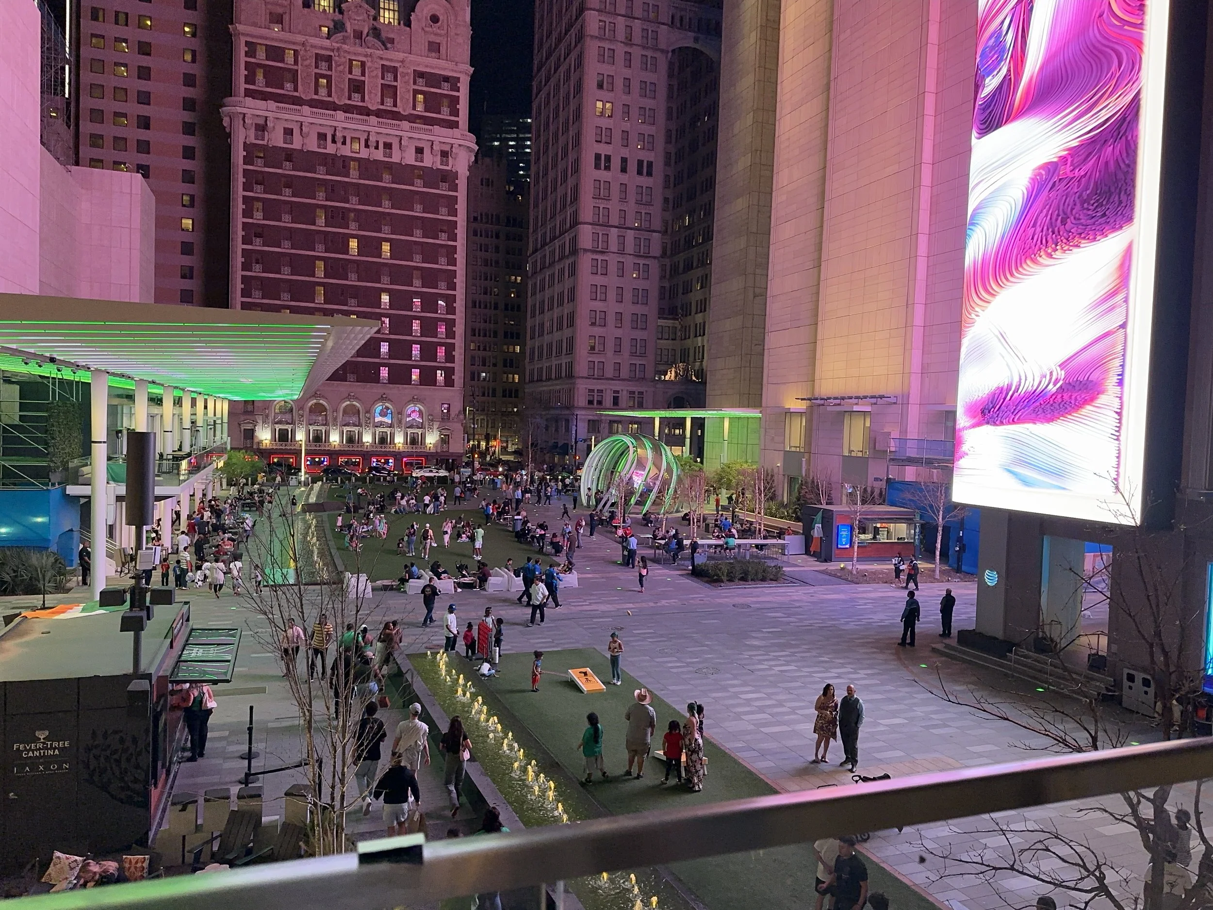 Night view of a city square with numerous people walking and gathering, surrounded by tall buildings illuminated with colorful lights and digital billboards.