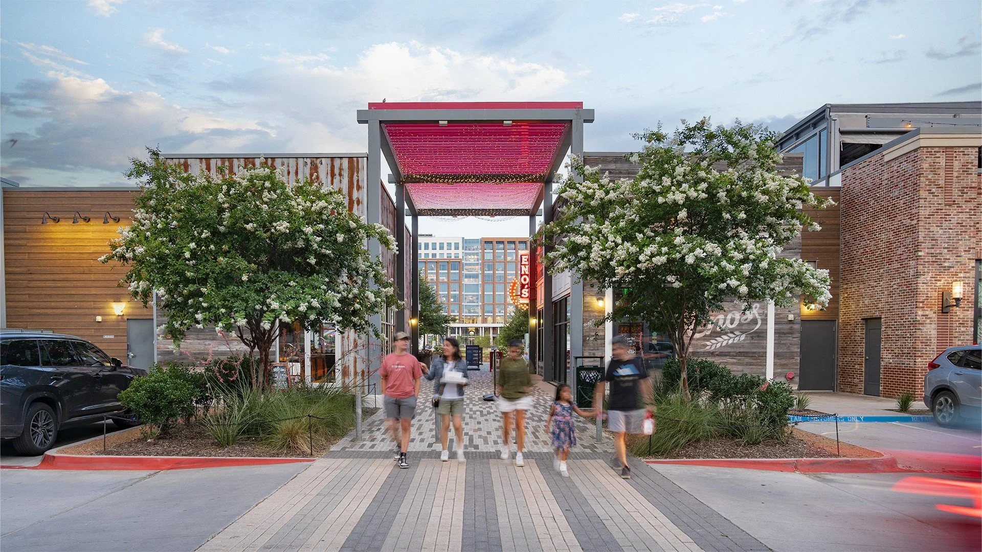 Scene of a shopping plaza entrance with people walking, two trees with white flowers, parked cars, and a building with a red and white sign reading 'Eino's' in the background.