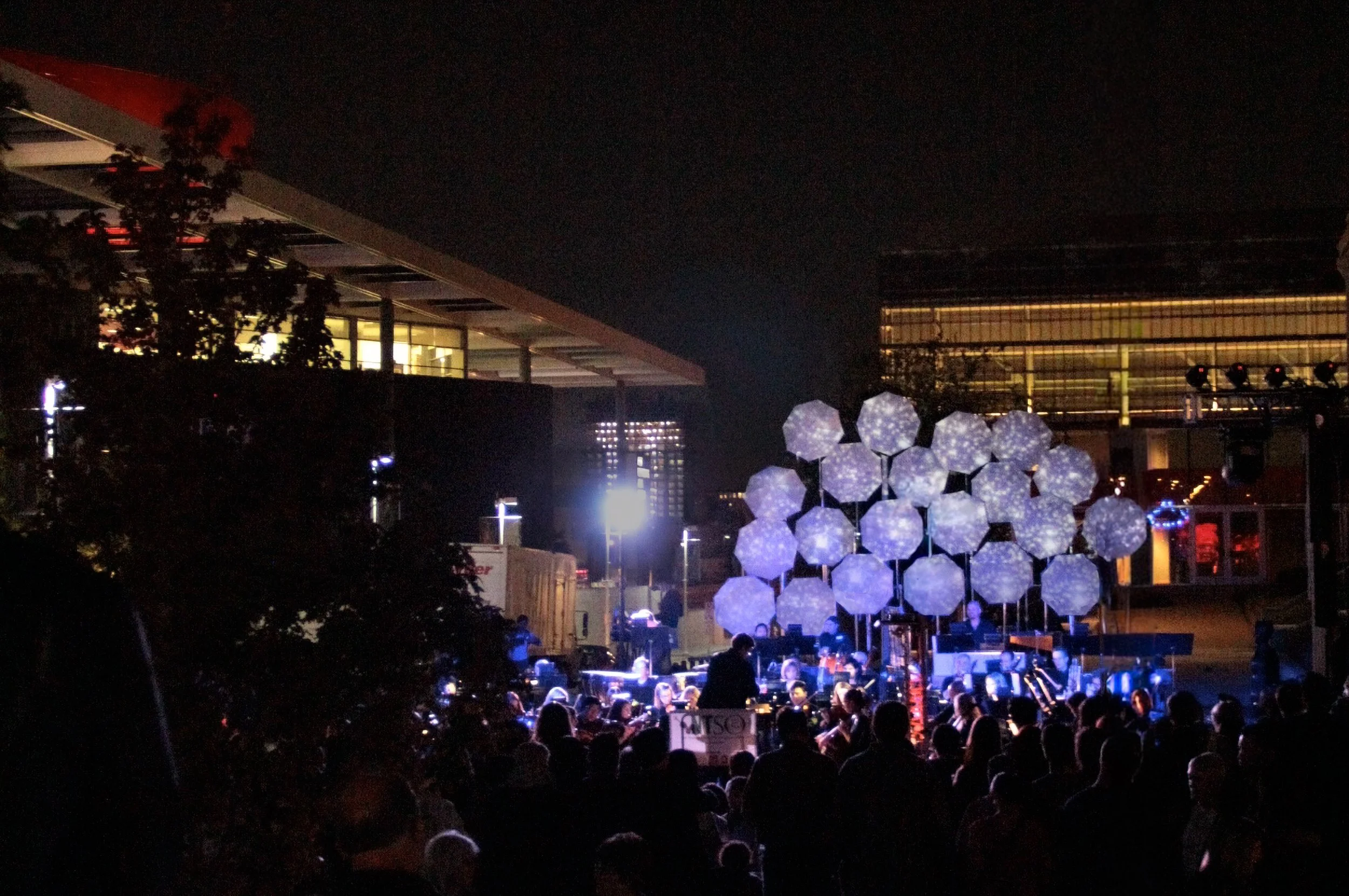 Nighttime outdoor concert with a large audience, illuminated stage, and decorative spherical light installation