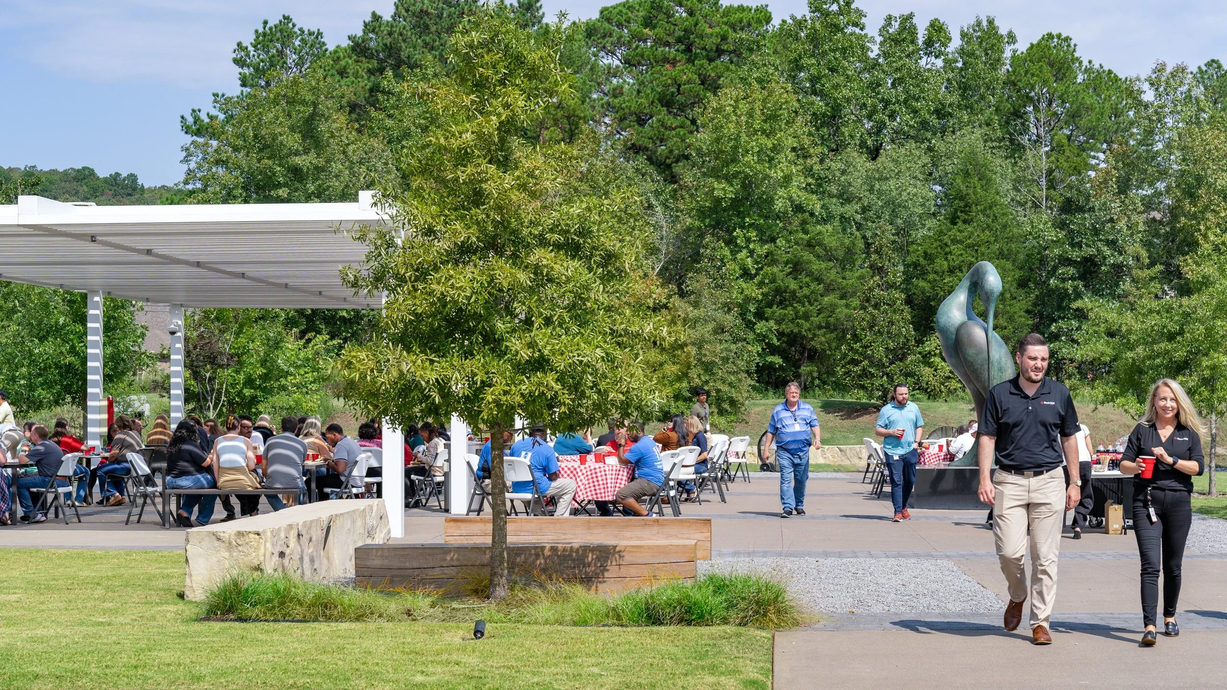 People dining outdoors at picnic tables with red and white checkered tablecloths in a park, with trees and a modern sculpture in the background.