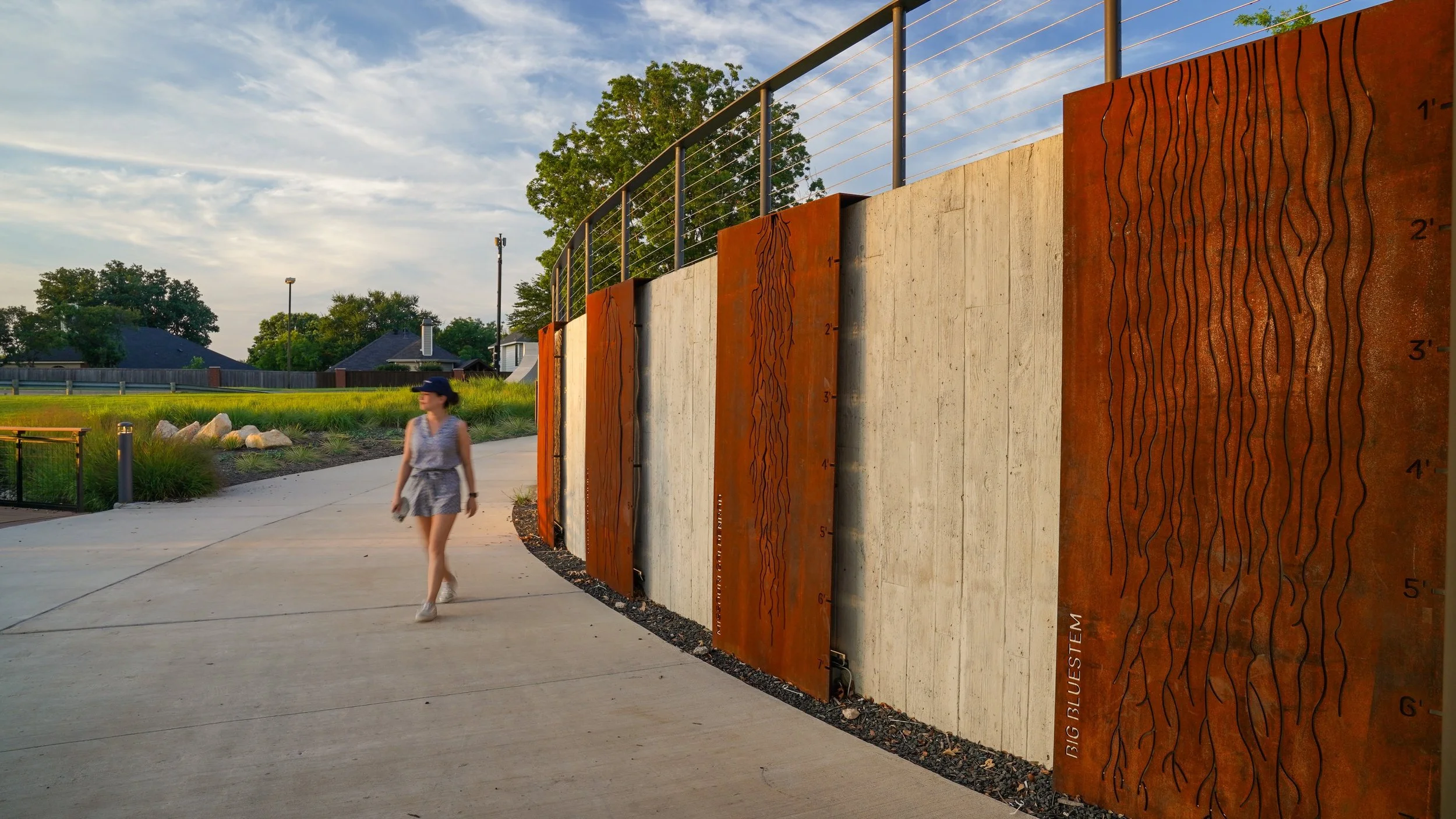 A woman walking on a paved path beside a decorative concrete and rusted steel wall with water flow design, in a park-like setting during sunset.