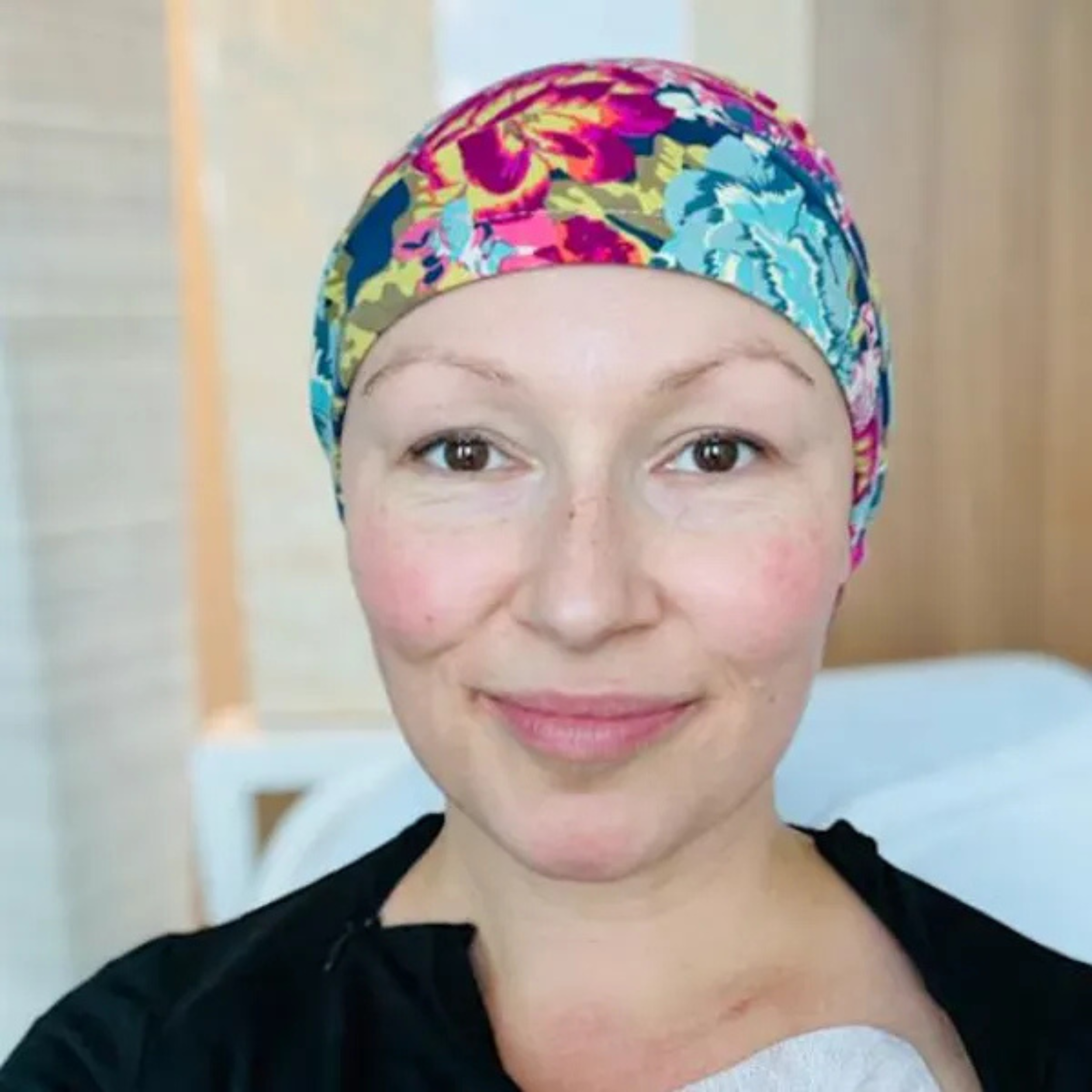A woman with light skin and brown eyes, smiling, wearing a colorful floral headscarf in a hospital room.