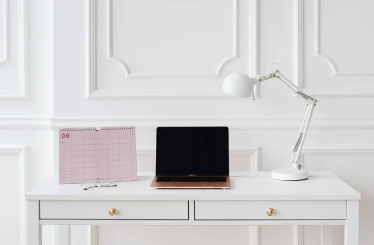 White desk against a white wall with decorative molding, holding a pink desk calendar, a closed black laptop, and a white adjustable desk lamp.