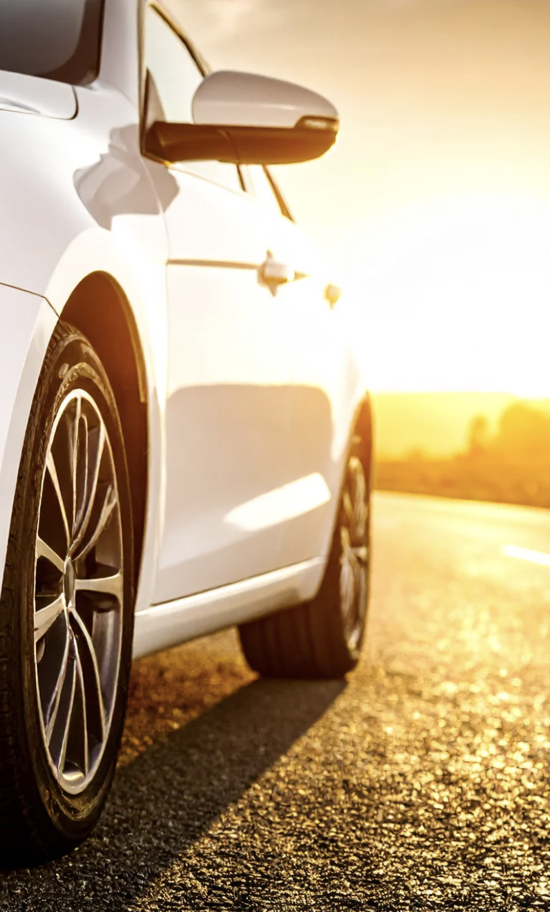Close-up of a white car parked on an empty road at sunset, focusing on the front wheel and side mirror.