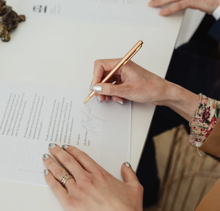 Person signing a document with a gold pen at a white desk, wearing a floral bracelet and a ring.