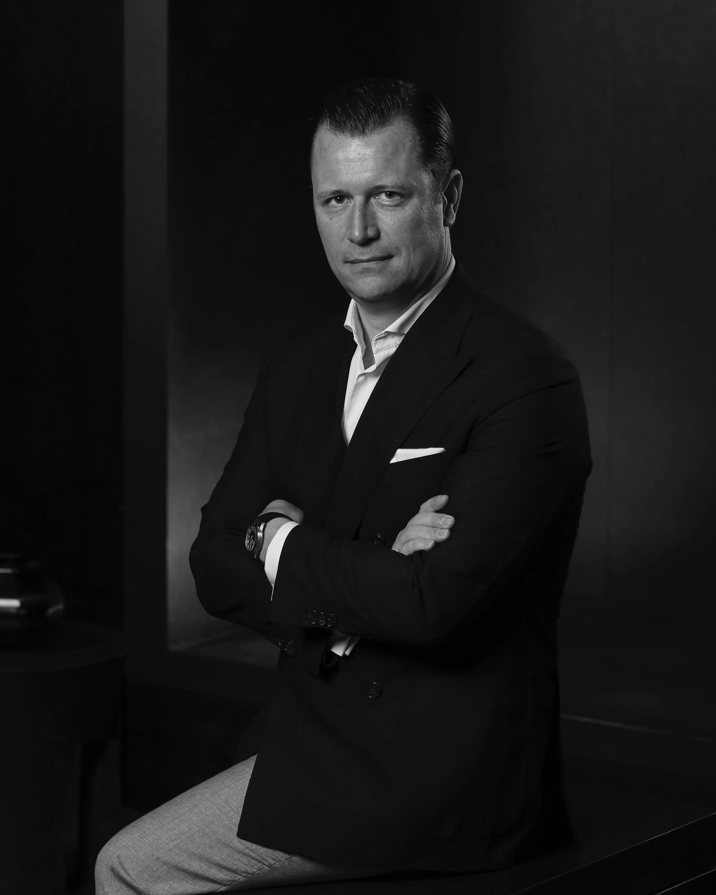 Black-and-white portrait of a man in a suit with his arms crossed, looking confidently at the camera.