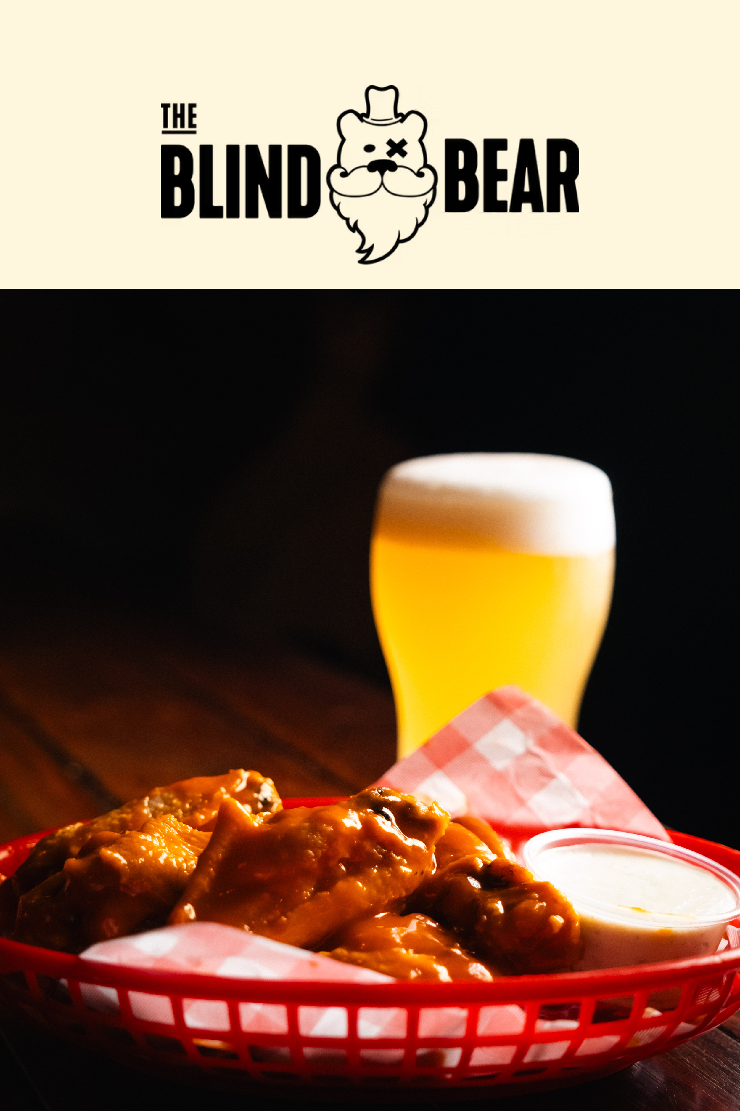 Basket of buffalo chicken wings with a side of ranch dressing, a glass of beer, and the Blind Bear logo at the top.
