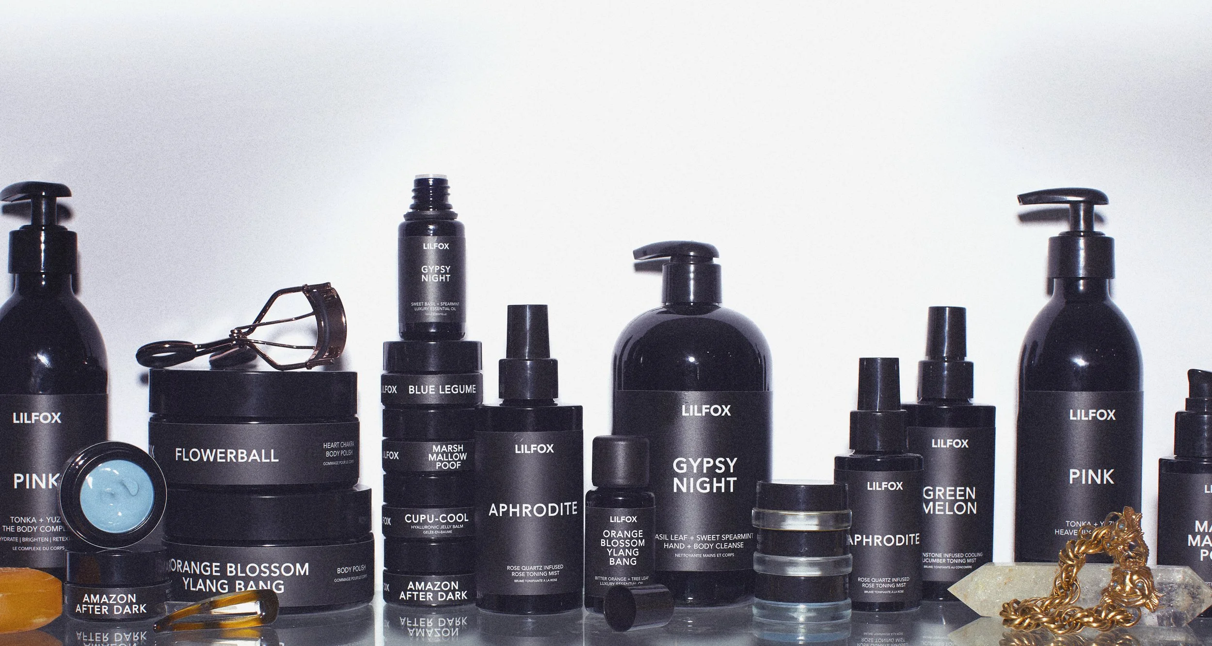 Arrangement of black cosmetic bottles and jars with white labels, some with pump dispensers, on a reflective surface with a plain white background.