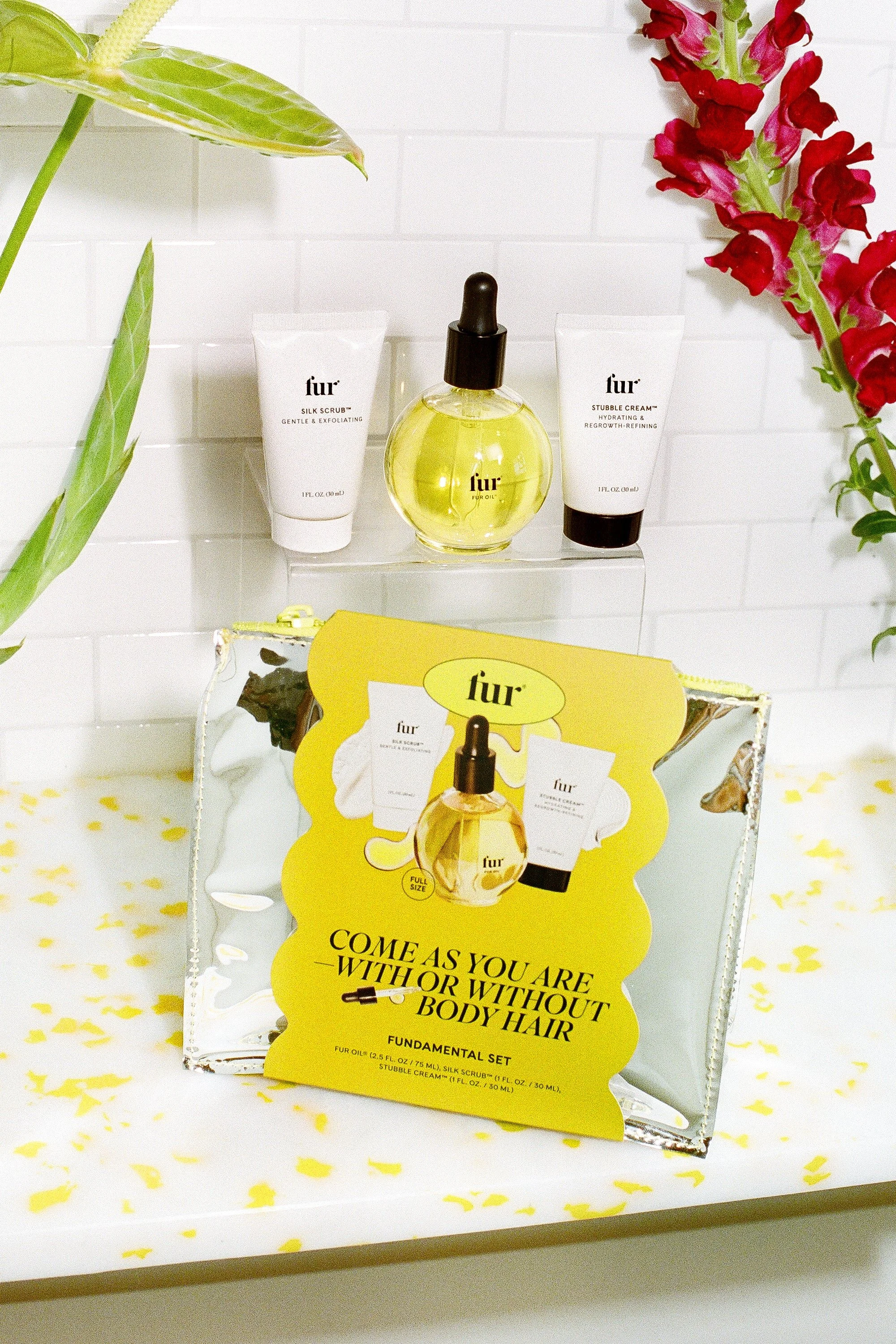 Fur skin care products including silk scrub and stubble cream, along with a dropper bottle of oil, displayed on a clear shelf with a decorative package in front, surrounded by green leaves and pink flowers.