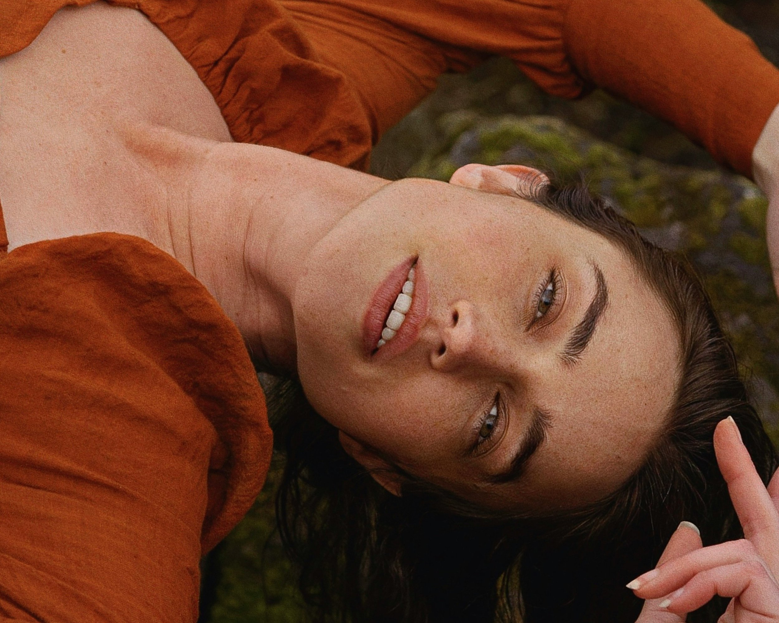 A woman with brown hair lies outdoors on mossy ground, wearing a rust-colored jacket, with a relaxed expression and slightly parted lips.