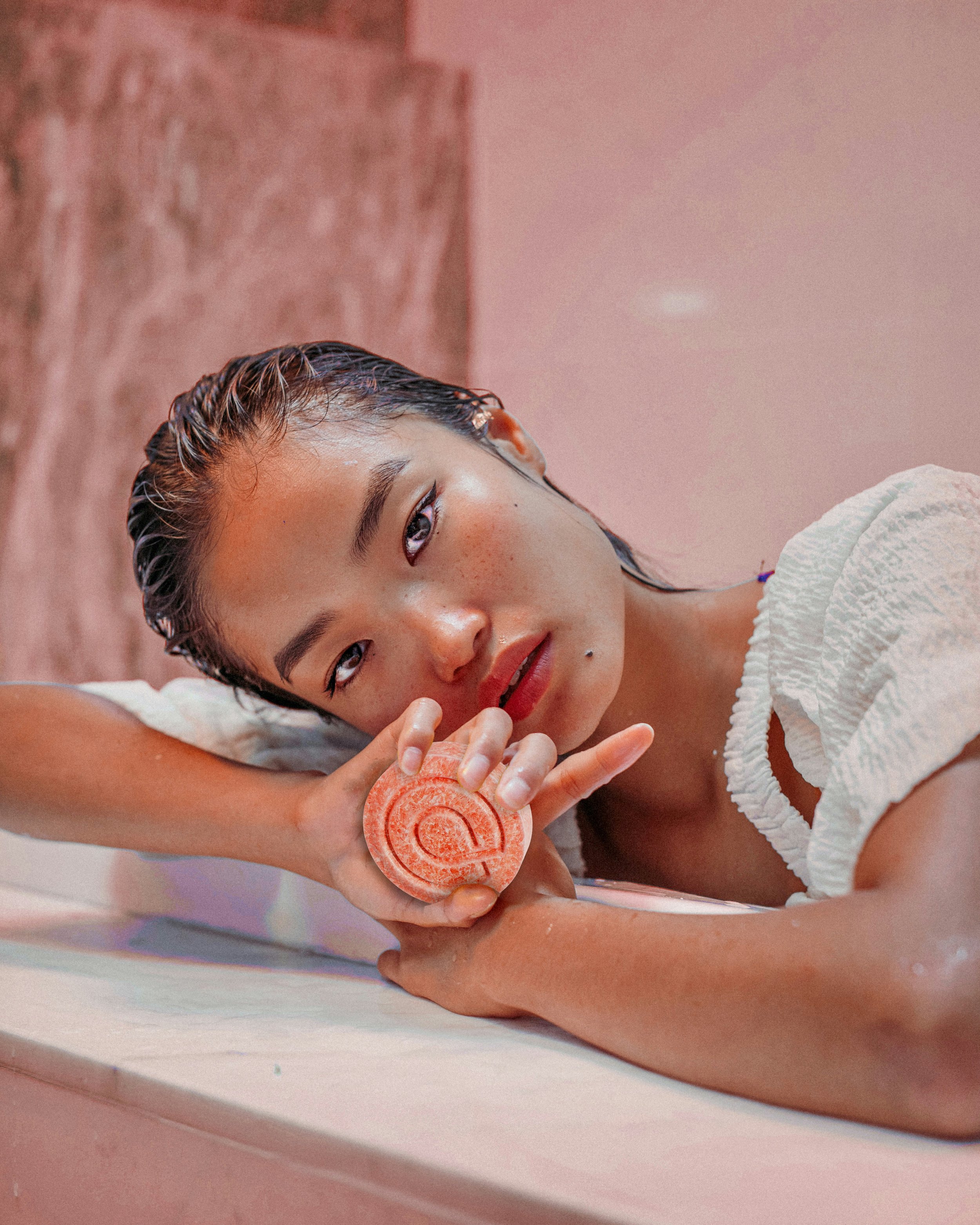 A young woman with wet hair, lying on her side with her head resting on her arm, holding an orange soap with a circular pattern close to her face. She has a relaxed expression and is wearing a white textured top, with a pink background and a pink marble wall behind her.