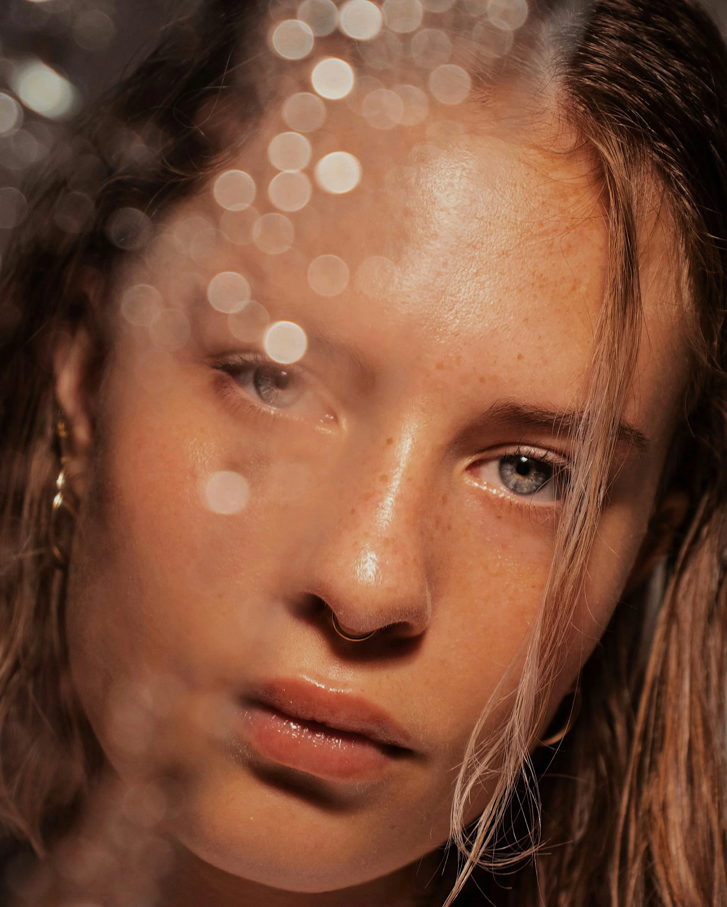 Close-up of a young woman with freckles, light skin, blue eyes, and wet hair, with bokeh lights in the foreground.