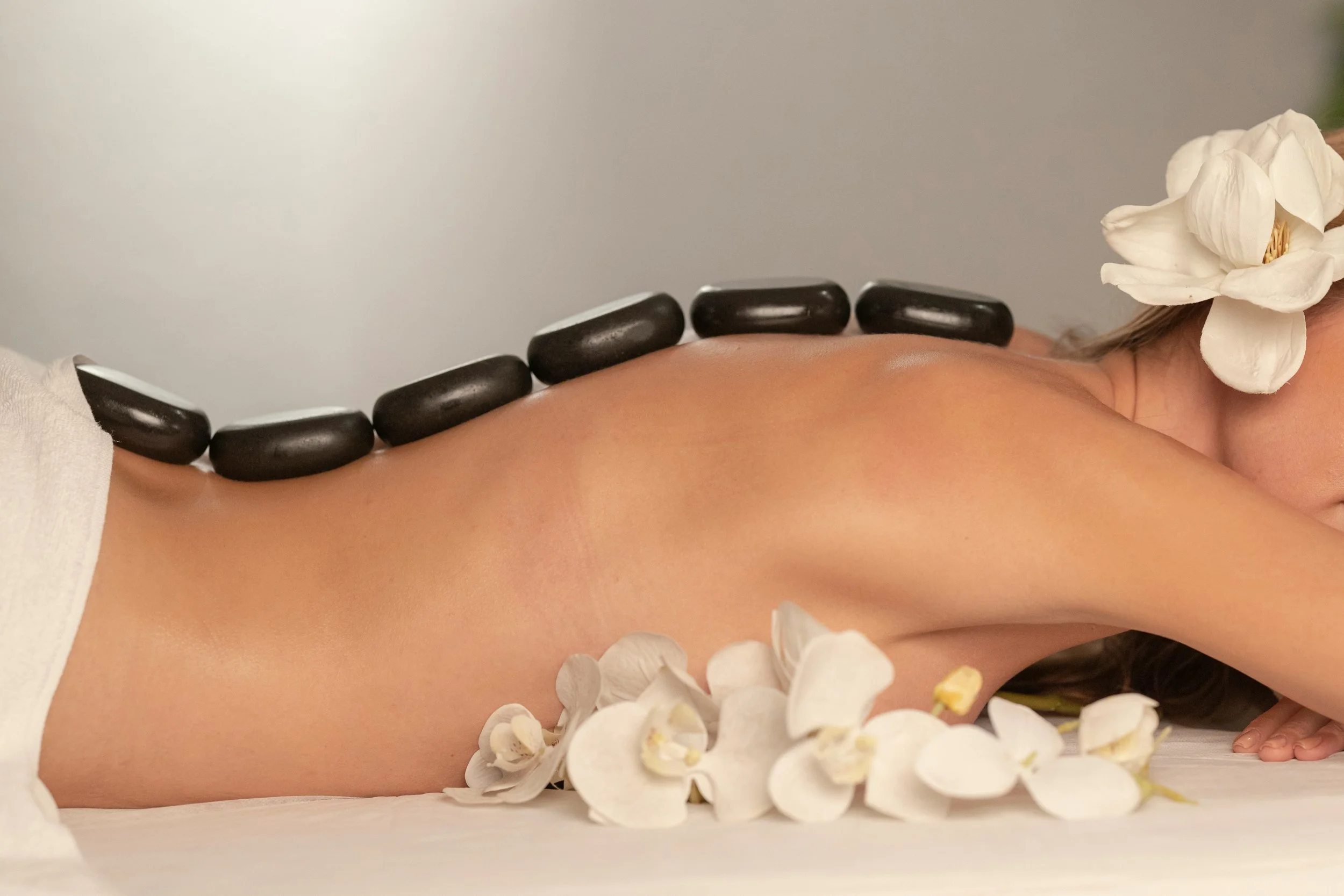 A woman lying face down with black hot stones placed along her upper back, surrounded by white orchid flowers.