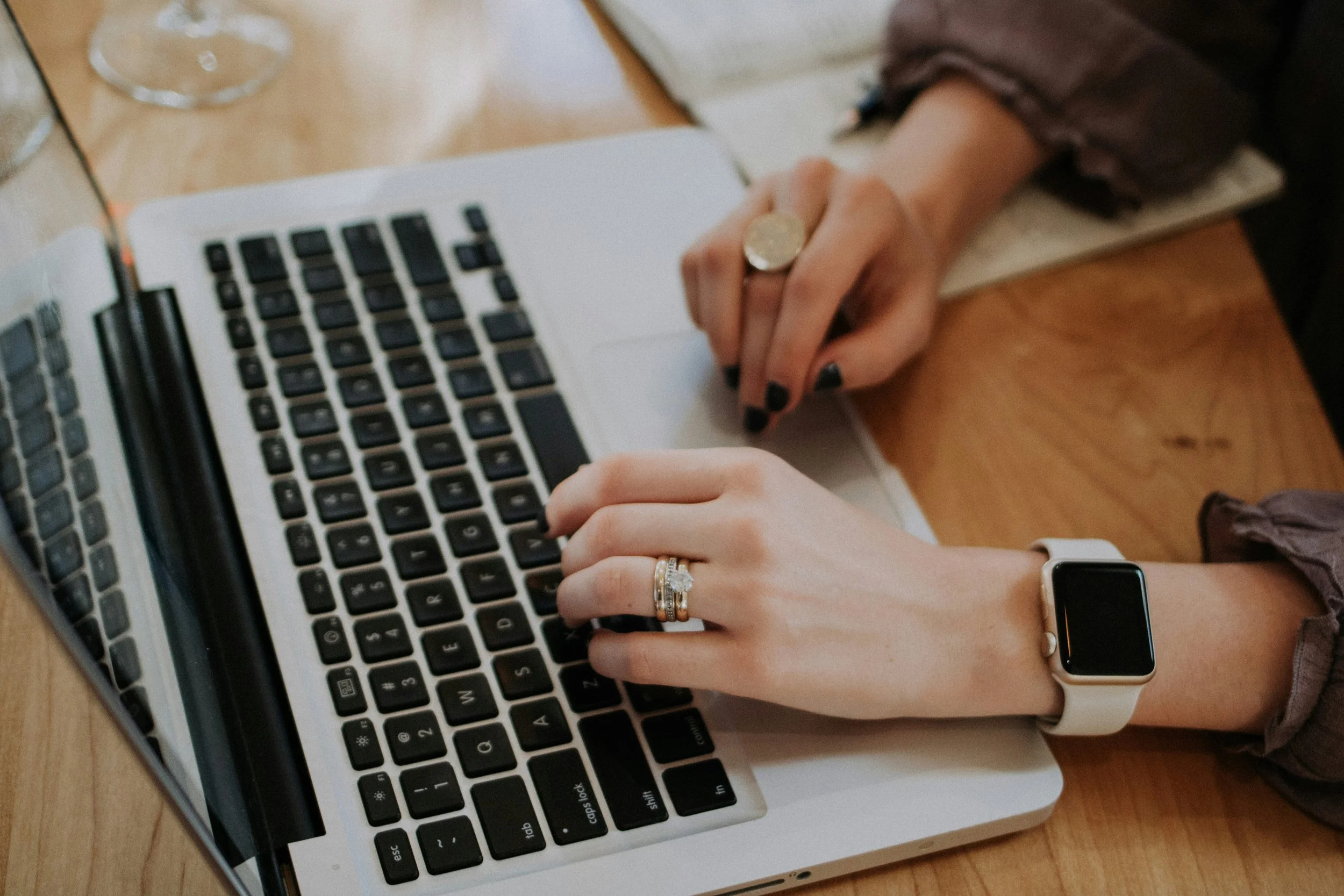 A person's hands typing on a laptop keyboard, with a wristwatch on the left wrist and rings on both hands. The person has black nail polish and is seated at a wooden table.