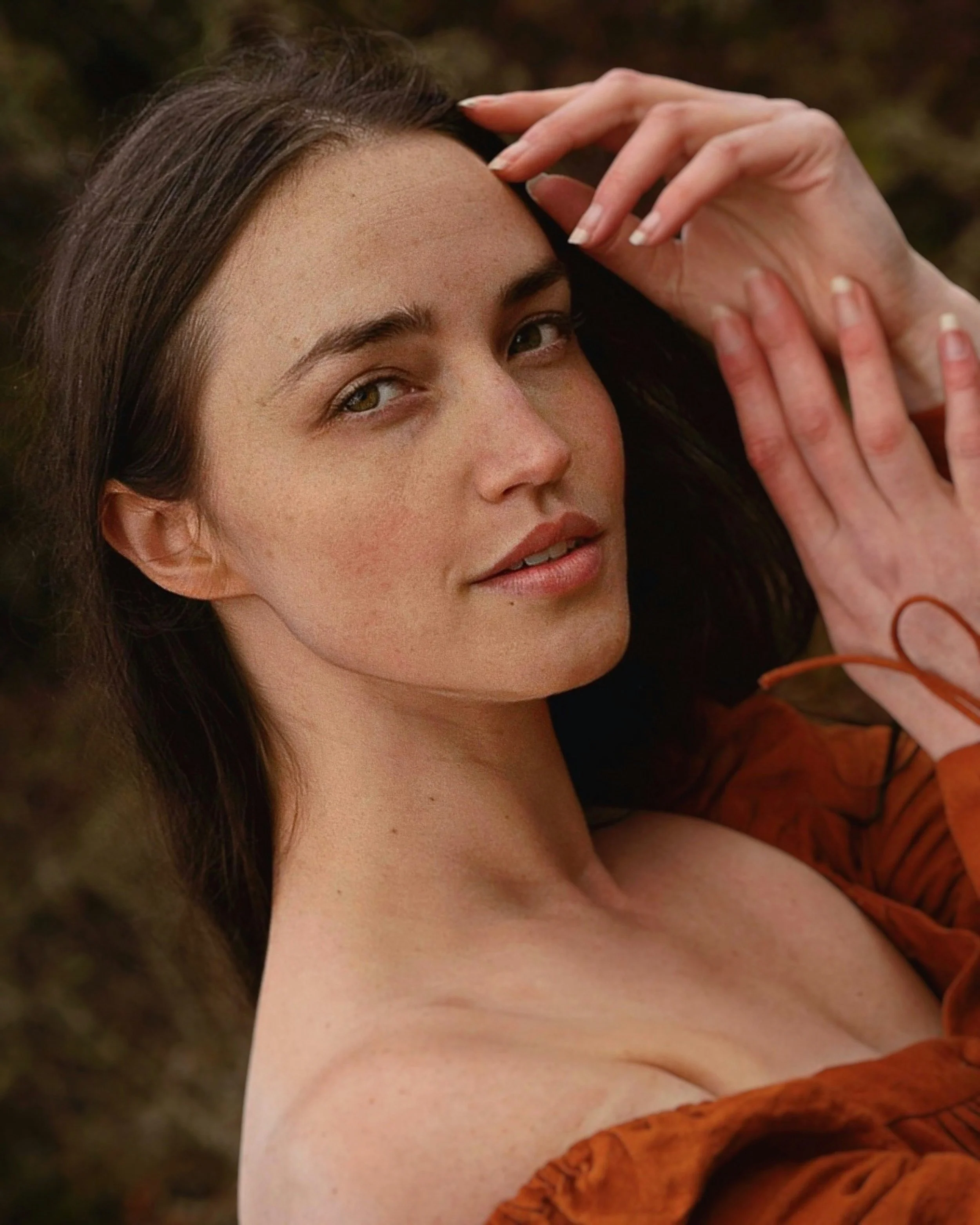 A young woman with dark hair and fair skin lies on her back, looking at the camera, with her right hand resting on her forehead and her left hand near her face. She is outdoors with a blurred natural background.