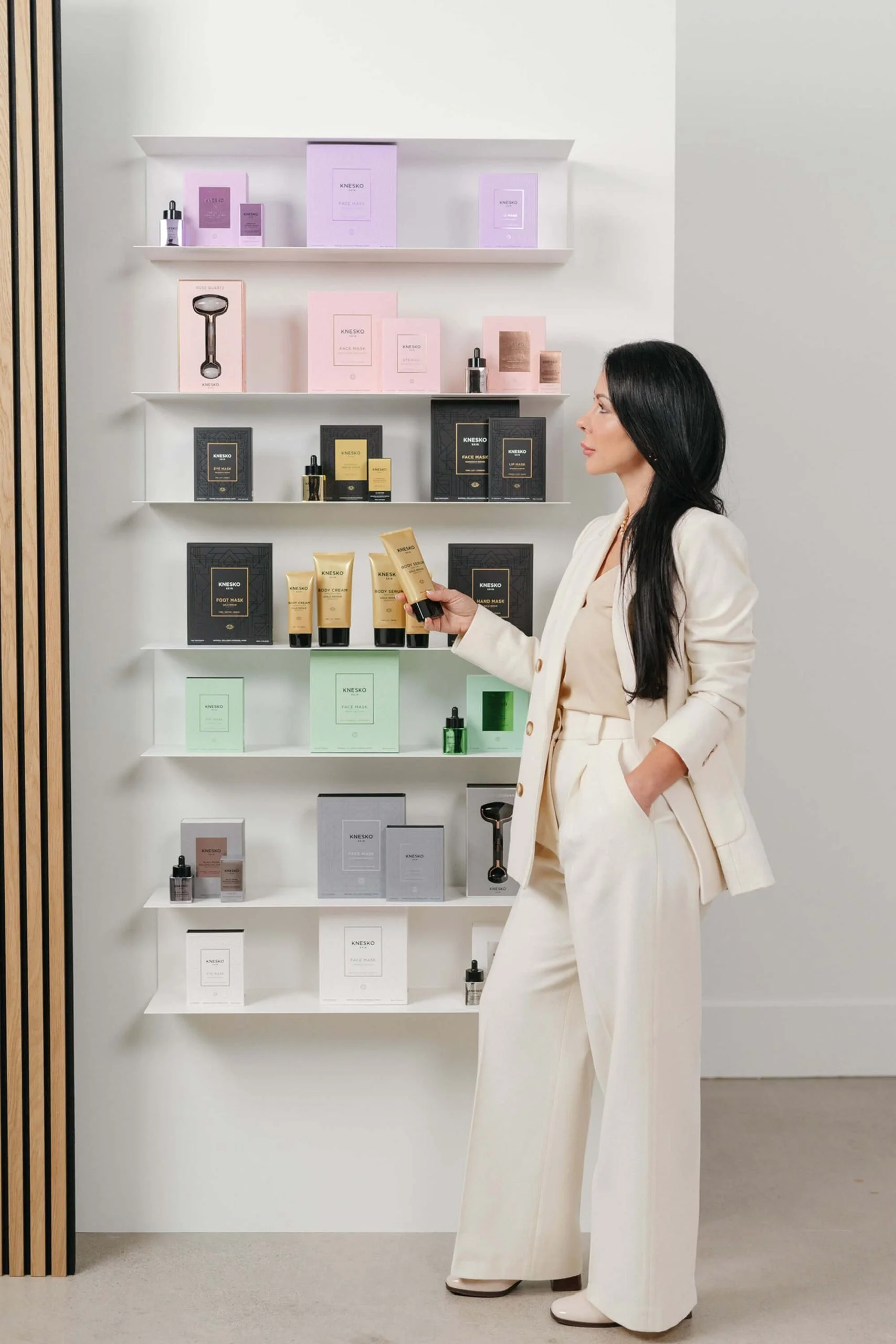 A woman with long black hair in a cream-colored suit shopping at a skincare and beauty product display shelf, with various boxed products of different colors.