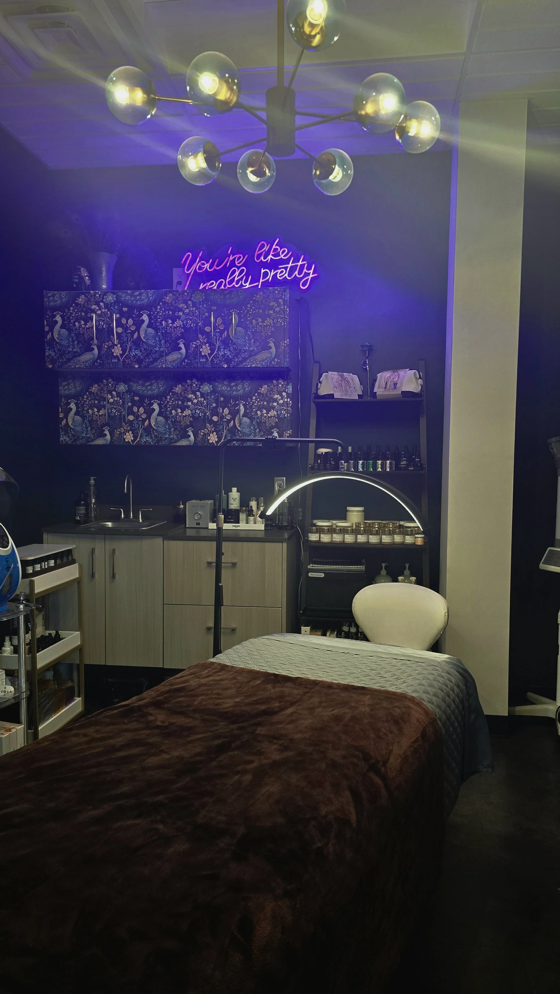 A cozy room with a massage bed covered in a brown blanket, a white chair, and a small kitchenette with skincare products and supplies. There is a neon sign on the wall that says 'You're like really pretty' and a modern chandelier with multiple bulbs hanging from the ceiling.