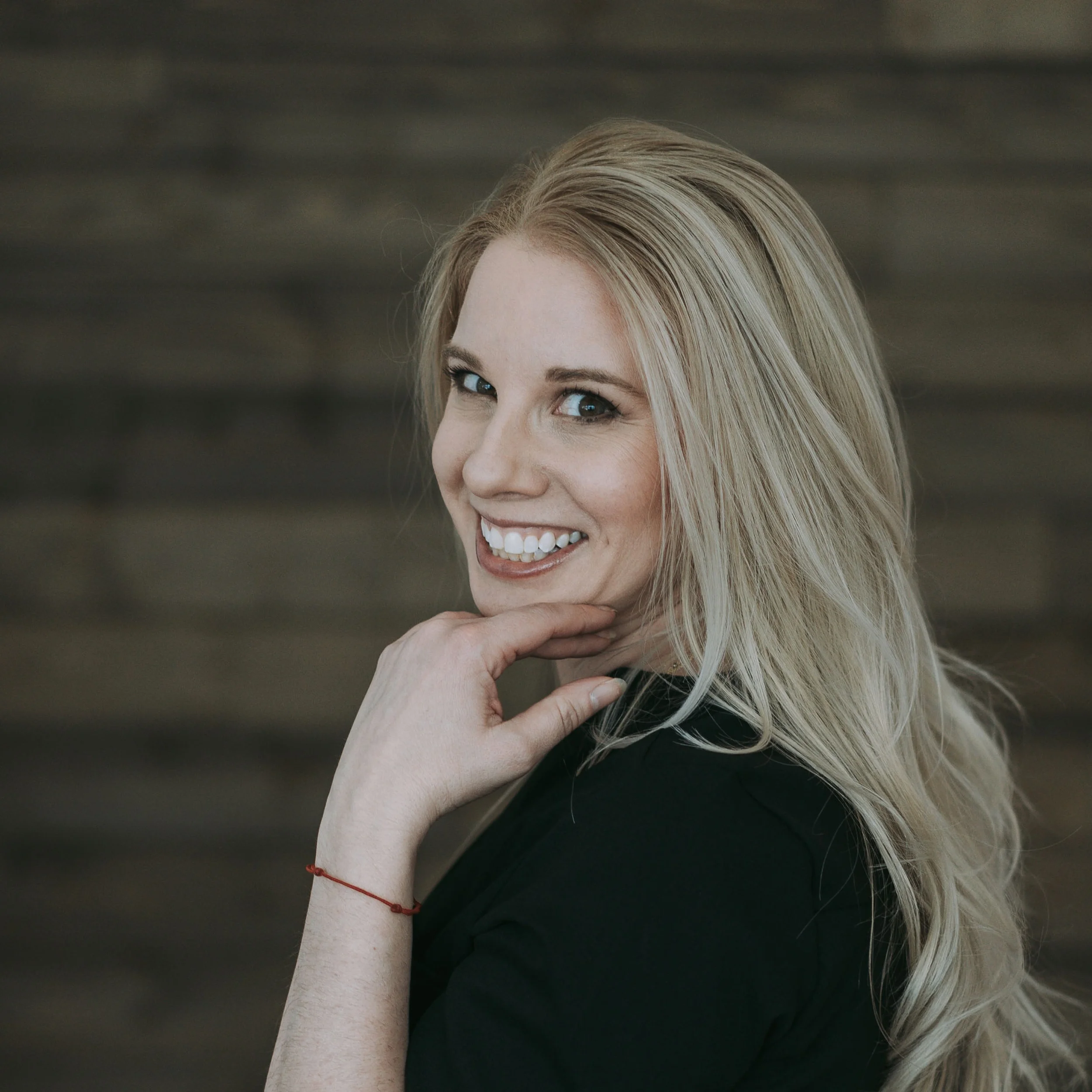 A blonde woman with long hair smiling and resting her chin on her hand, wearing a black top against a wooden background.
