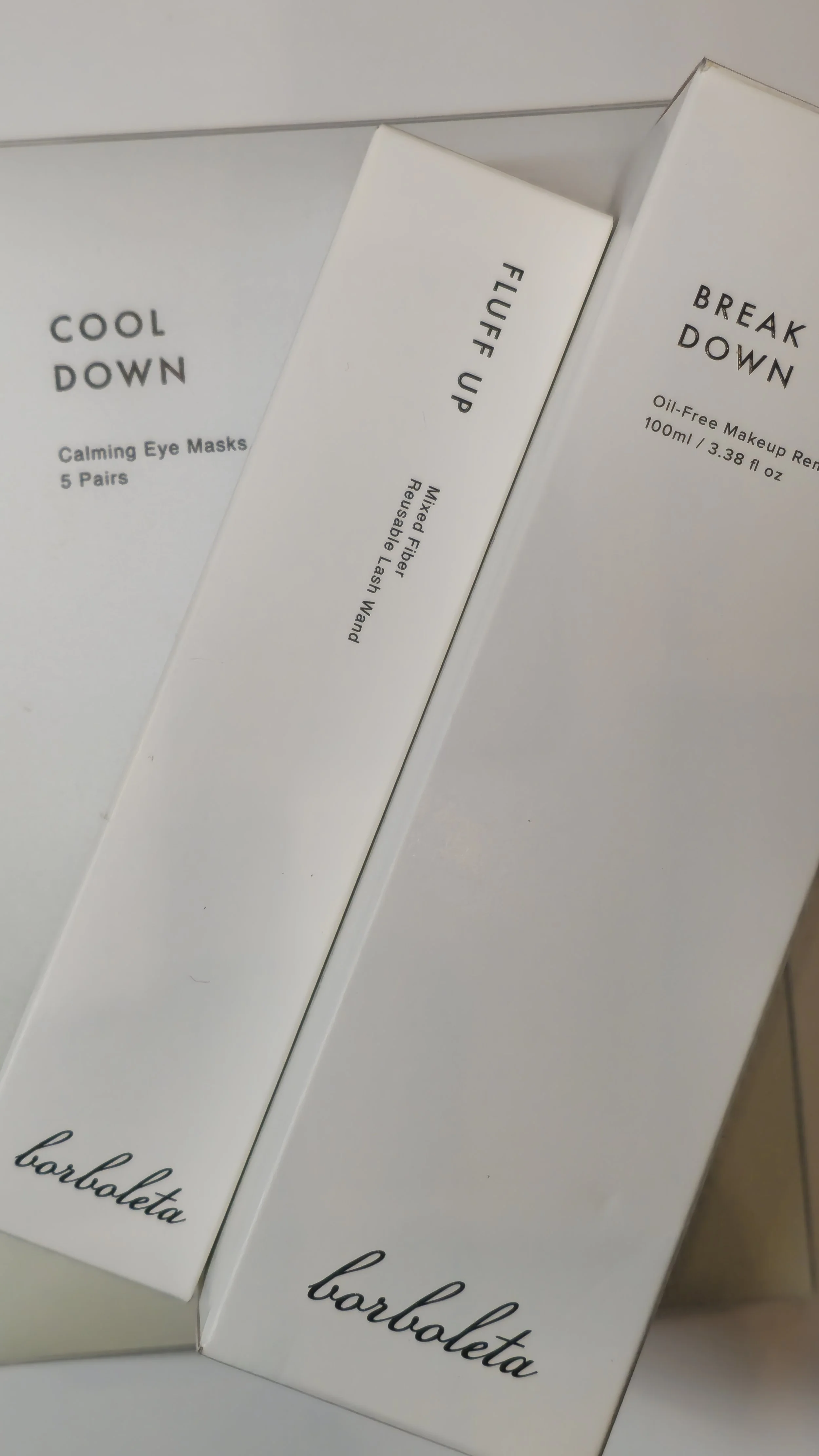 Three skincare product boxes from the brand barbaretta, arranged on a reflective surface. The products include 'Cool Down' calming eye masks, 'Fluff Up' micro fiber reusable lash wand, and 'Break Down' oil-free makeup remover.