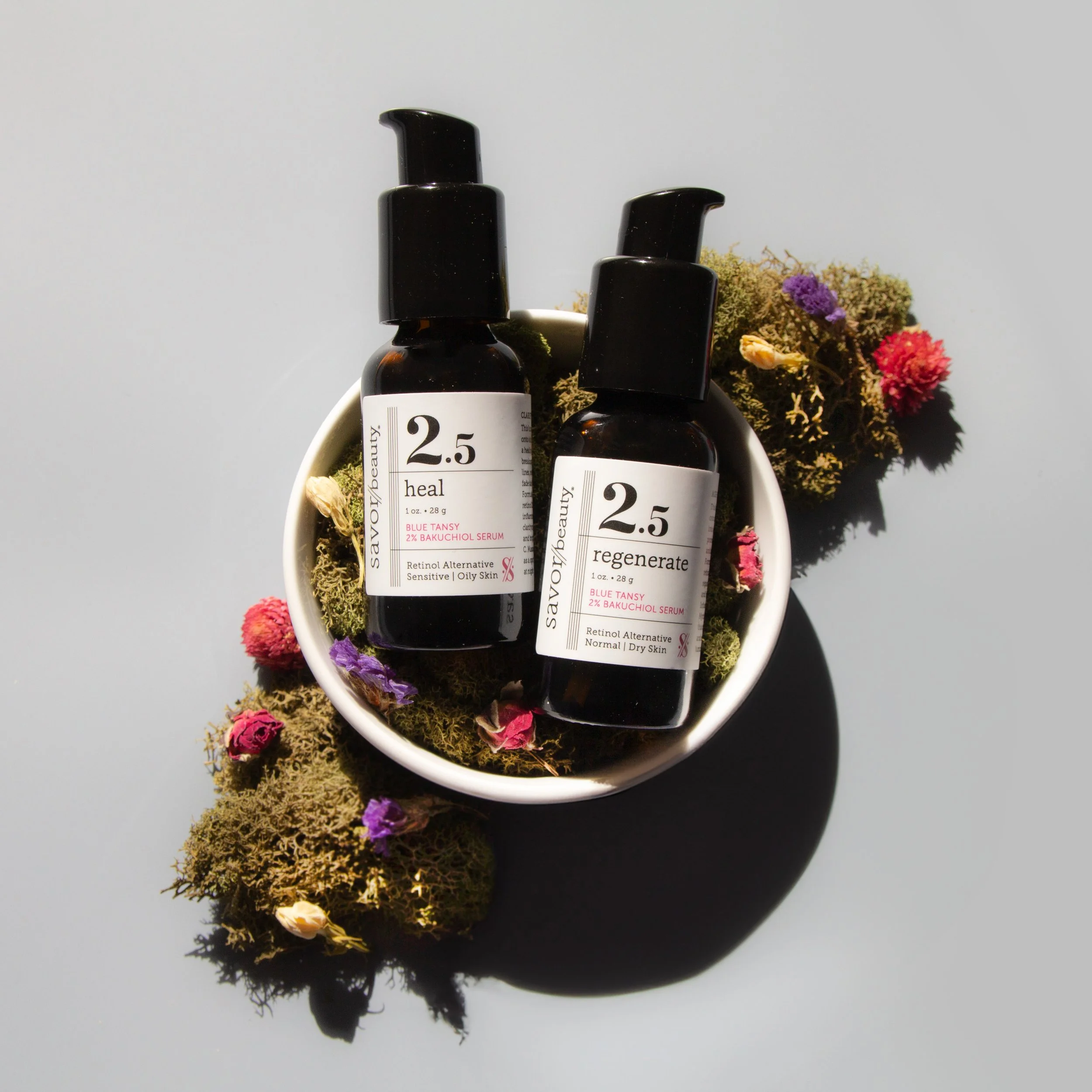 Two amber glass bottles of skincare serum labeled 'savior/beauty 2.5 heal' and 'savior/beauty 2.5 regenerate' are placed in a white bowl surrounded by moss and dried flowers on a light gray background.