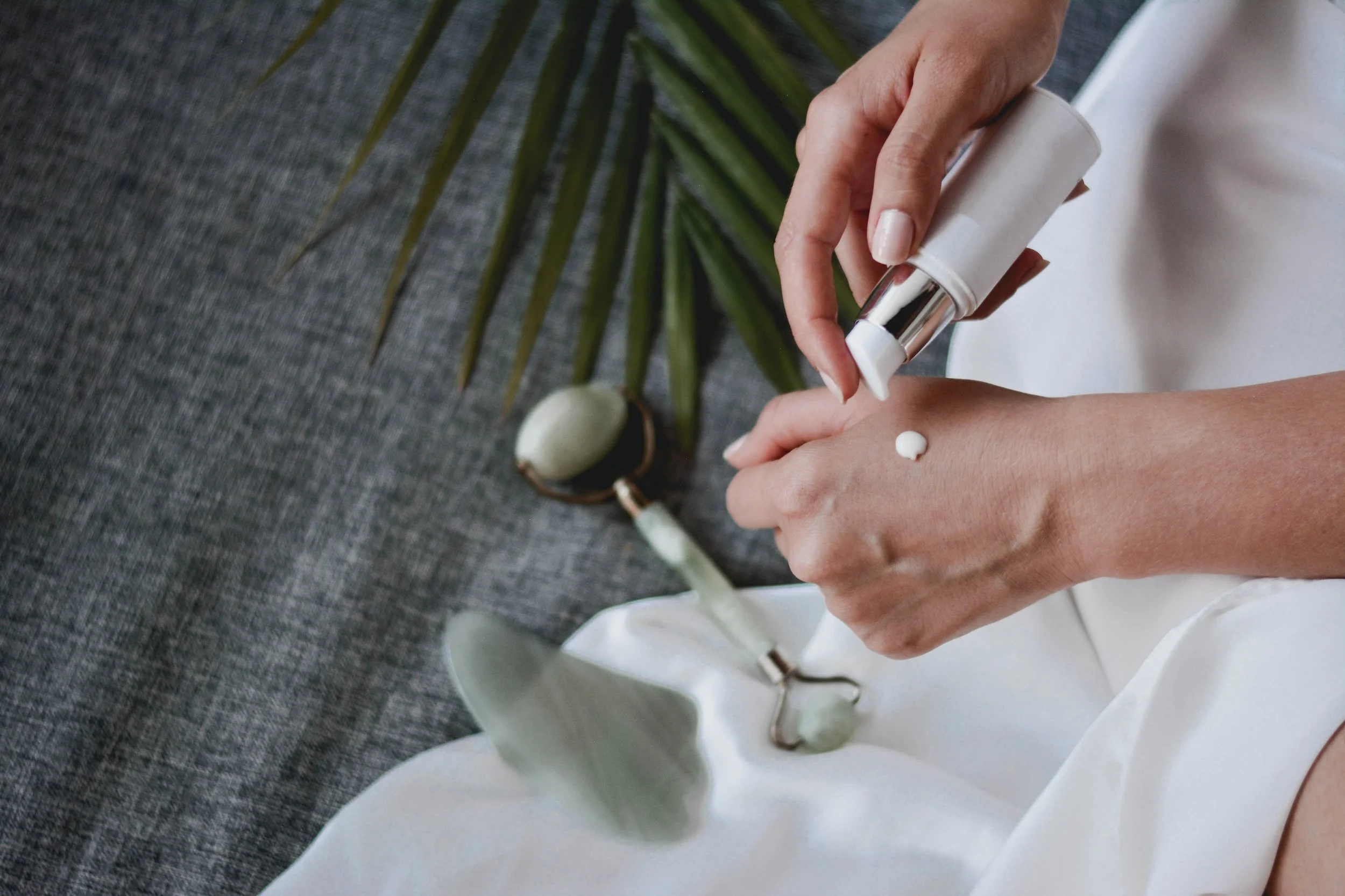 Person applying moisturizer to their hand from a pump bottle, with skincare tools and a palm leaf in the background.