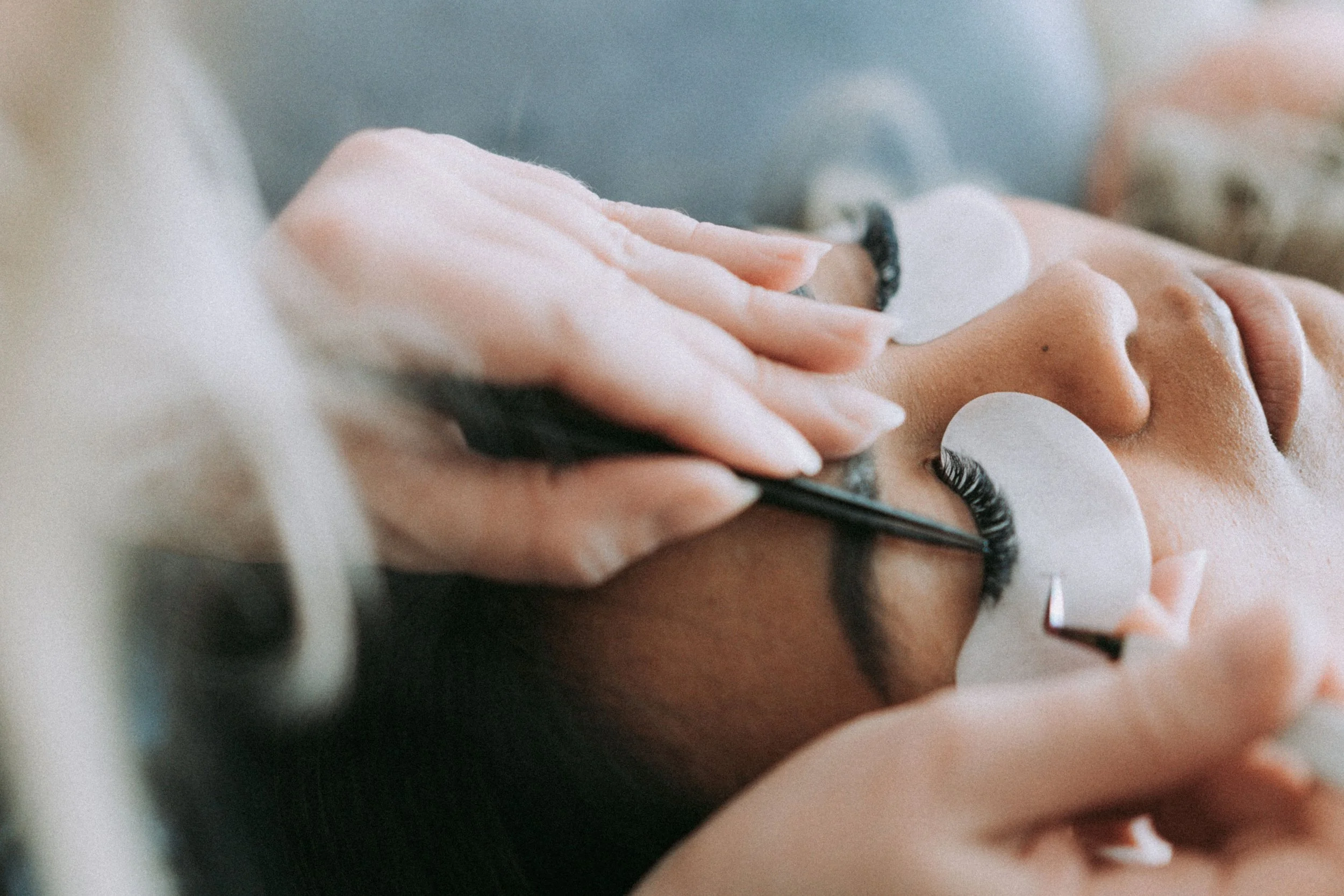 A woman lying down with her eyes closed, getting eyelash extensions applied by a technician using a thin tool.