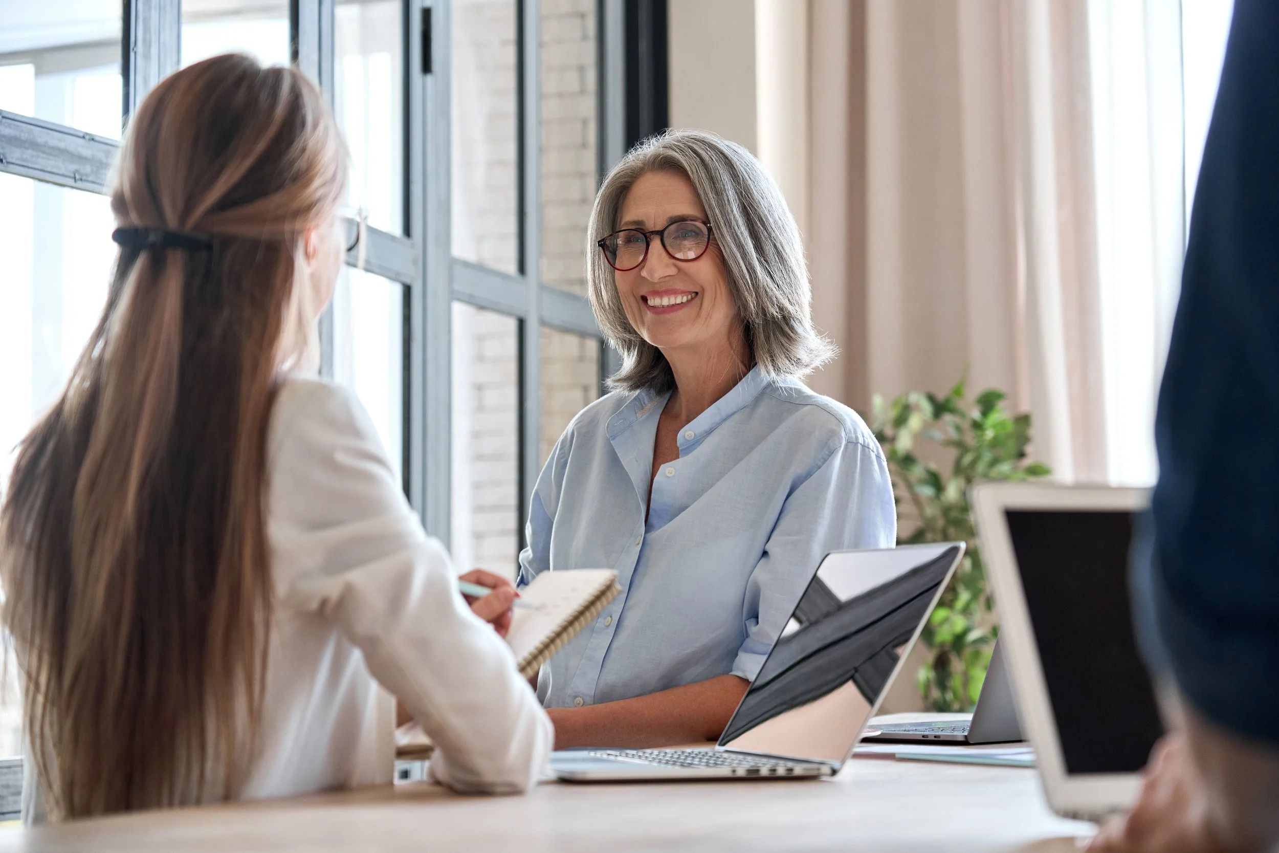 An older woman with gray hair and glasses smiling while talking to a young girl with long brown hair in a bright office space.