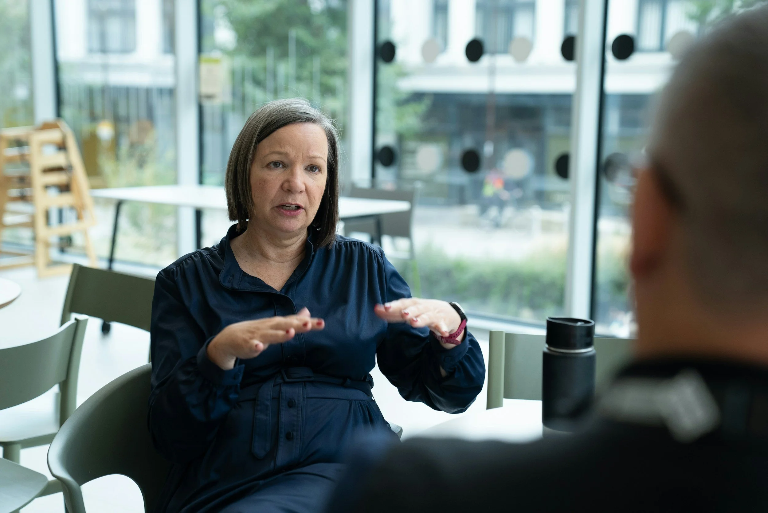 A woman with dark hair and a blue shirt gesturing with her hands during a conversation in a bright room with large windows and green trees outside.