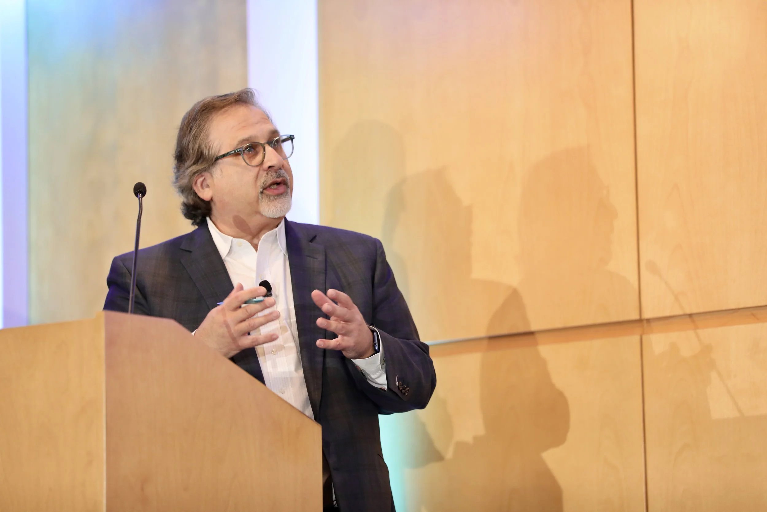 Geoffrey Fenton, with glasses wearing a suit, speaking at a podium during a presentation.