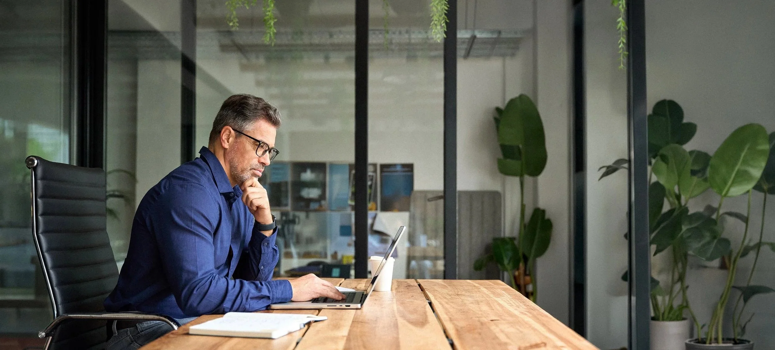 A man in glasses and a blue shirt sitting at a wooden desk, working on a laptop in a modern office with glass walls and large green plants in the background.