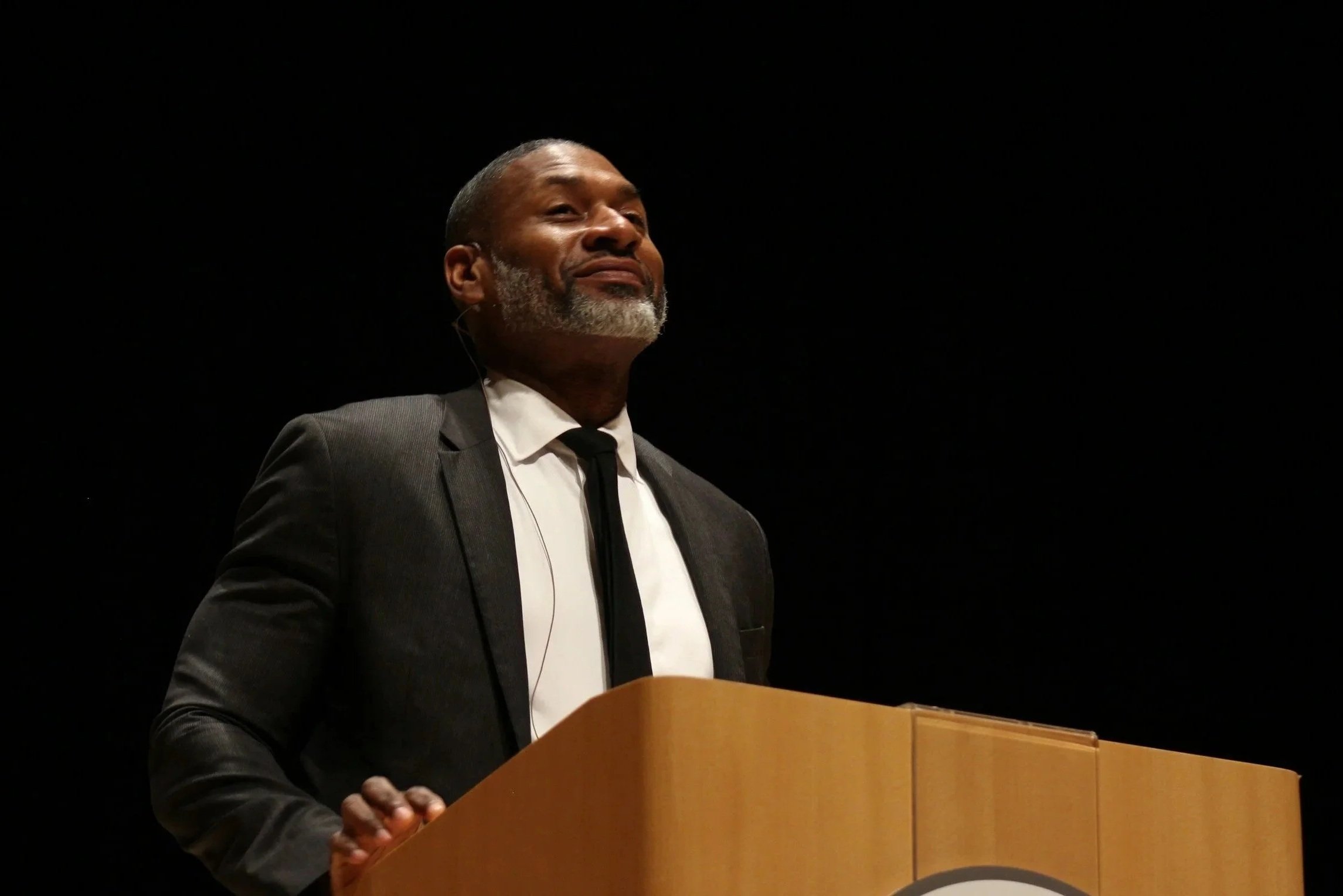 An African American man with a beard and gray hair, wearing a suit and tie, stands behind a wooden podium, speaking or presenting in a dark, likely auditorium setting.