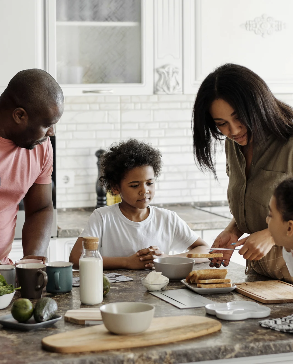 Family preparing breakfast together in kitchen, with a woman handing a toast to a child while man watches, surrounded by food items and kitchenware.