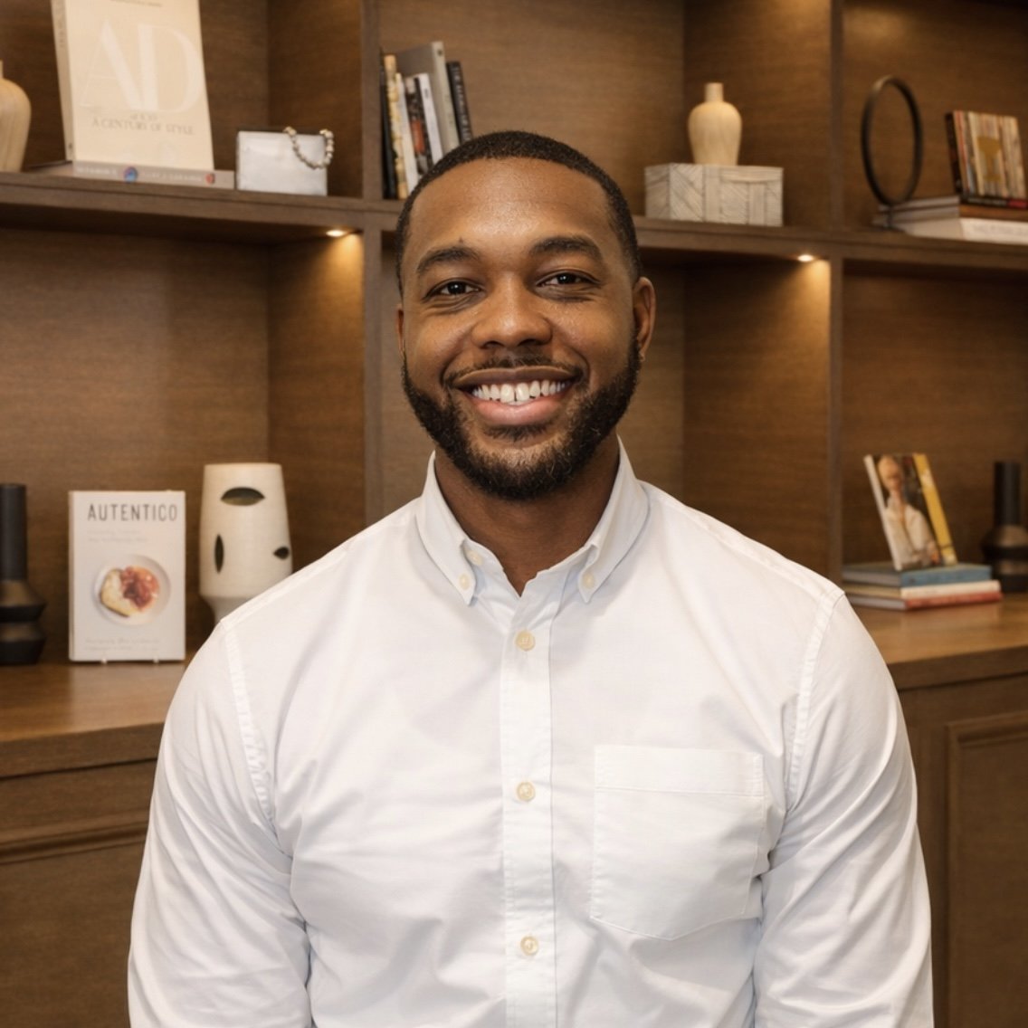 Smiling man wearing a white button-up shirt in front of a wooden bookshelf with books and decorative objects.