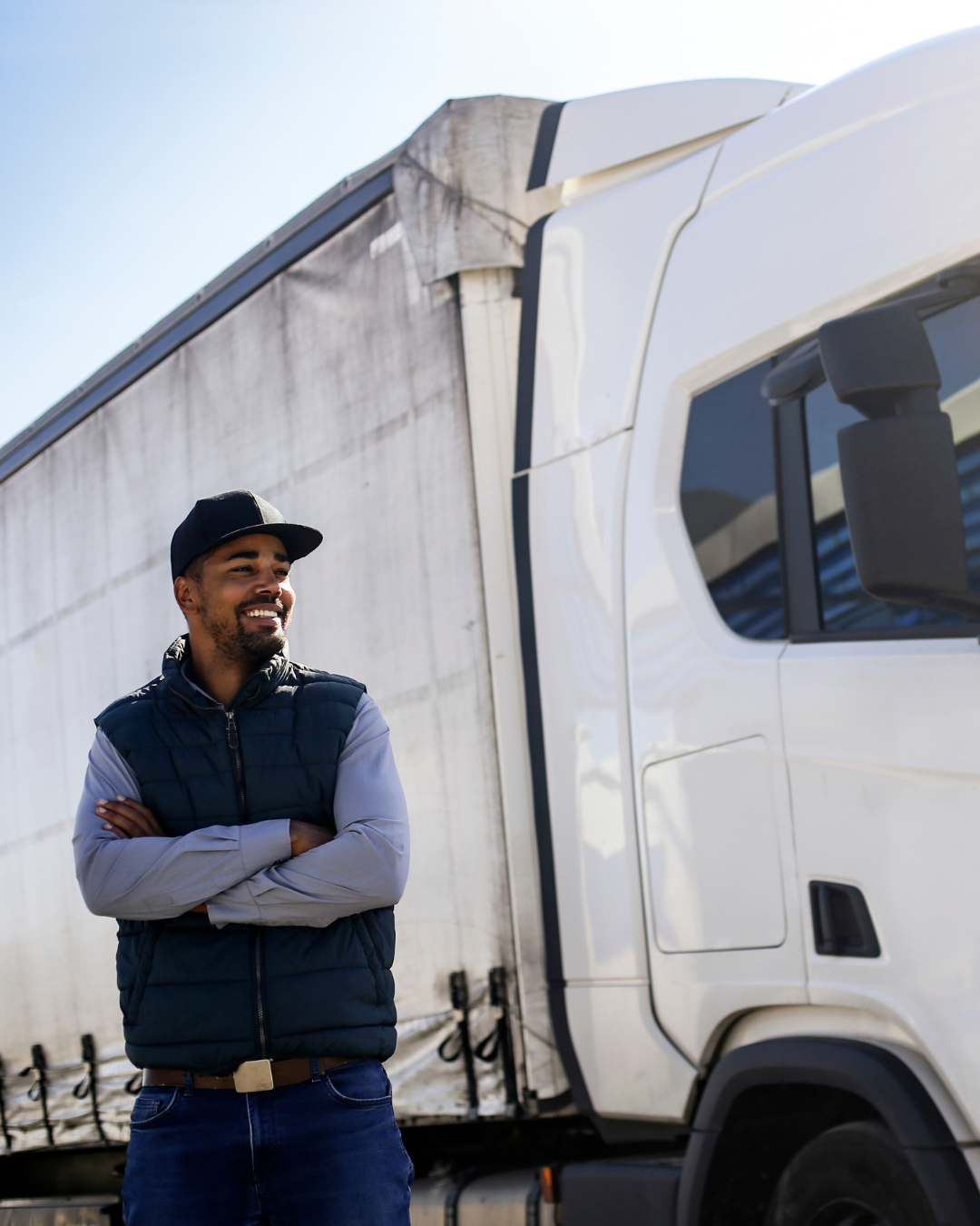 A man in a black cap and navy vest leaning against a white semi-truck with a smile.