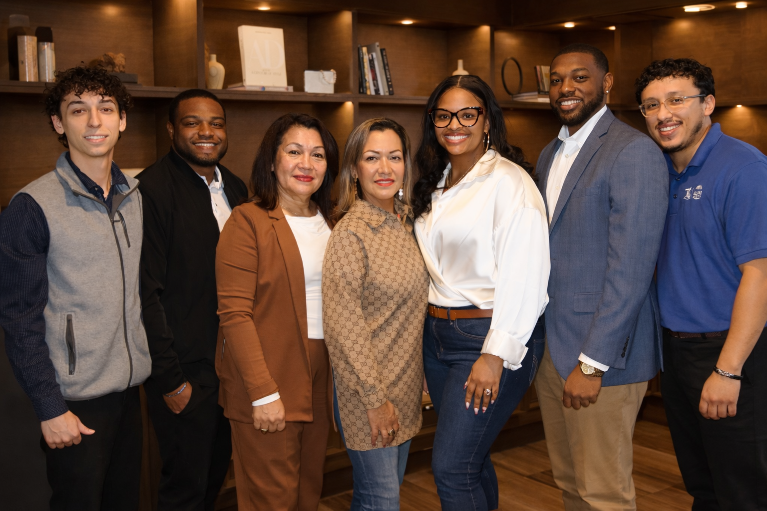 Group of eight diverse adults standing together indoors, smiling at the camera.
